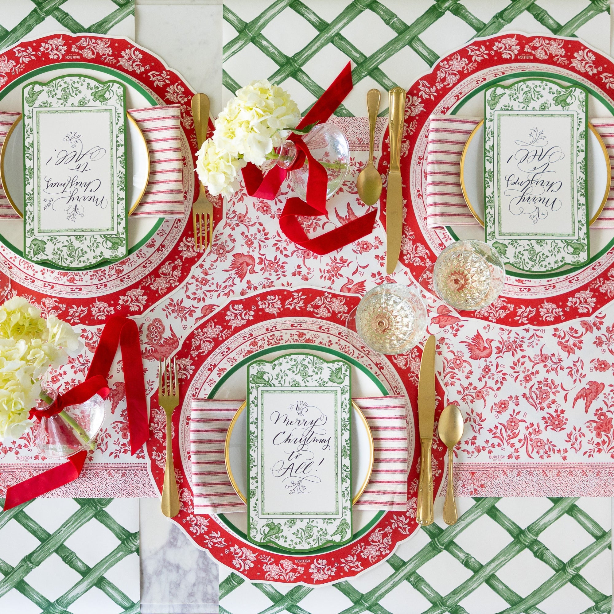 A table for three is set featuring the Die-cut Red Regal Placemat, dinner plates with a red-and-white striped napkin in between, Green Regal Peacock Table Accent reading "Merry Christmas to All!" atop, gold flatware, Green Lattice Runner rolled out vertically underneath and Red Burleigh Peacock Runner rolled out horizontally underneath.