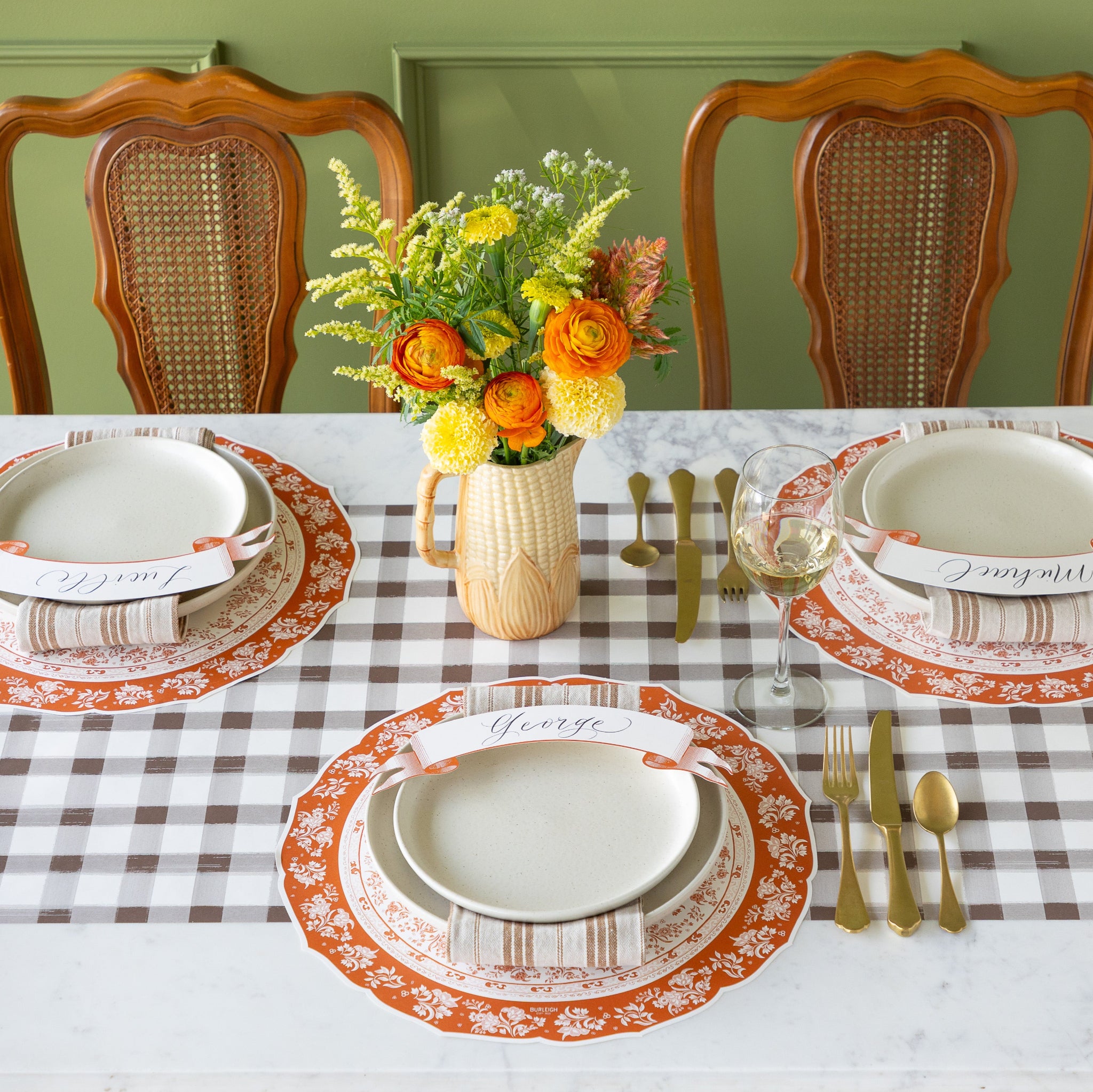 A table setting for three with flowers in a corn-shaped vase, featuring the Die-cut Pumpkin Regal Placemat, off-white dinnerware, napkins, Classic Orange Banner Table Accents and gold flatware on the Brown Painted Check Runner.