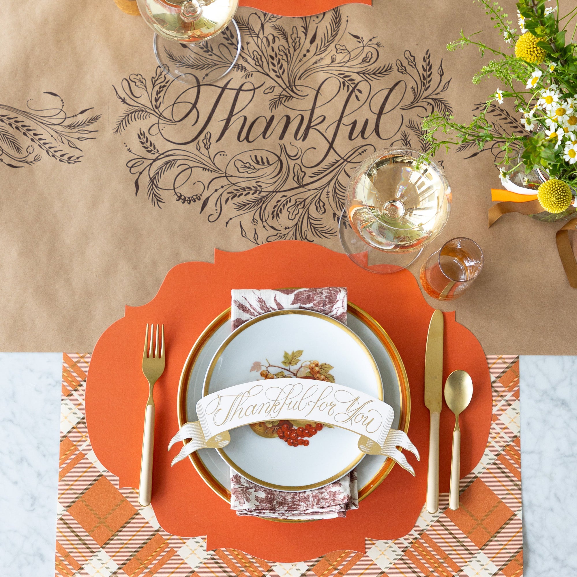 A fall table setting featuring the Orange French Frame Placemat with vintage plates, a floral napkin and Thankful for you Banner Table Accent atop, tan flatware, and  a wine glass on the Kraft Thankful Runner.