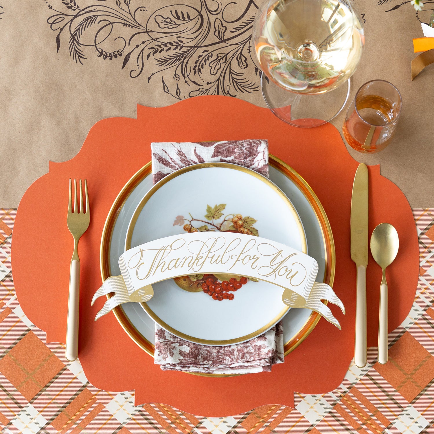 A fall table setting featuring the Orange French Frame Placemat with vintage plates, a floral napkin and Thankful for you Banner Table Accent atop, tan flatware, and  a wine glass on the Kraft Thankful Runner.