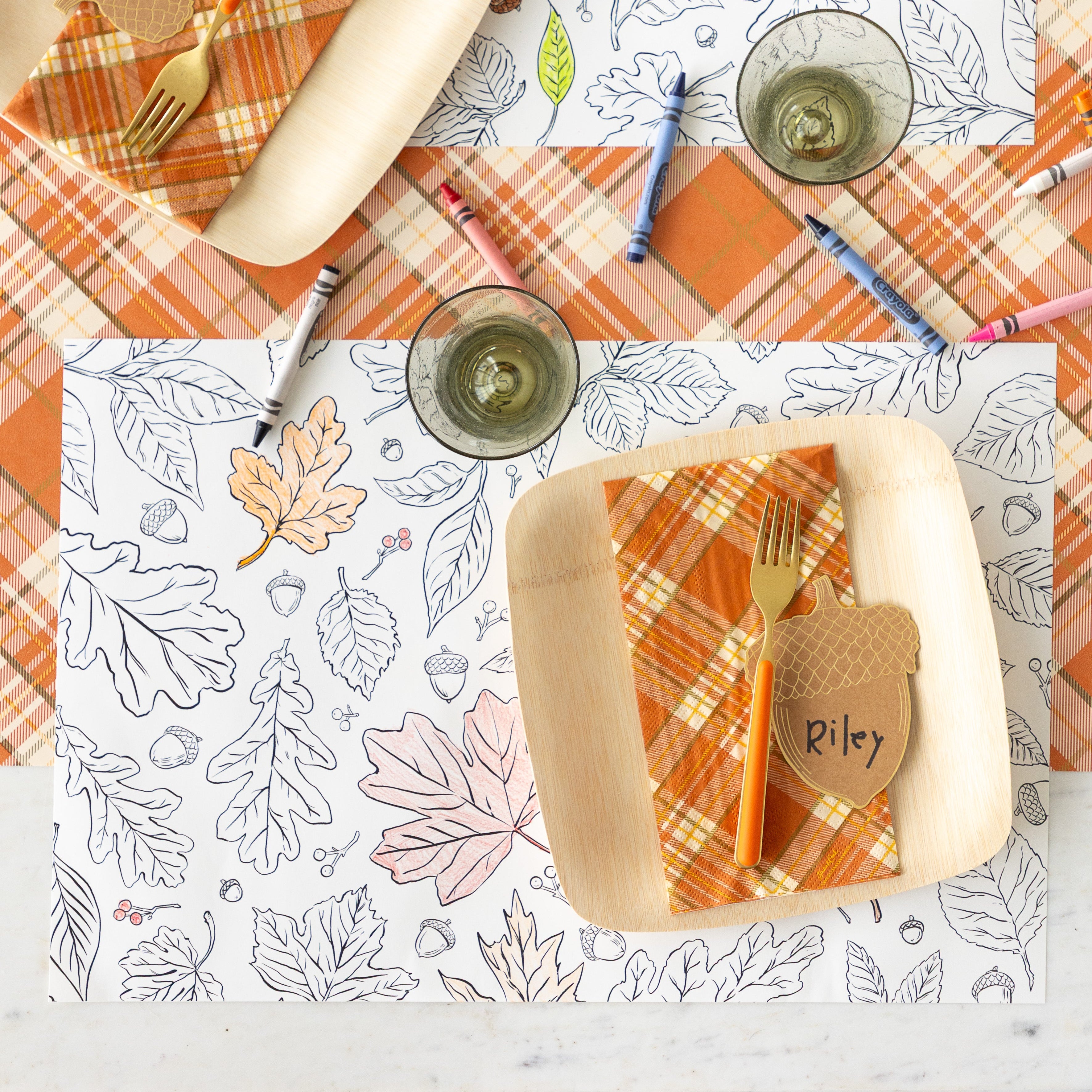 A place setting featuring the Coloring Leaves Placemat partially colored in, a paper plate with the Orange Plaid Guest Napkin, orange fork and Acorn Place Card reading &quot;Riley&quot; on top and scattered crayons on the Orange Plaid Runner.