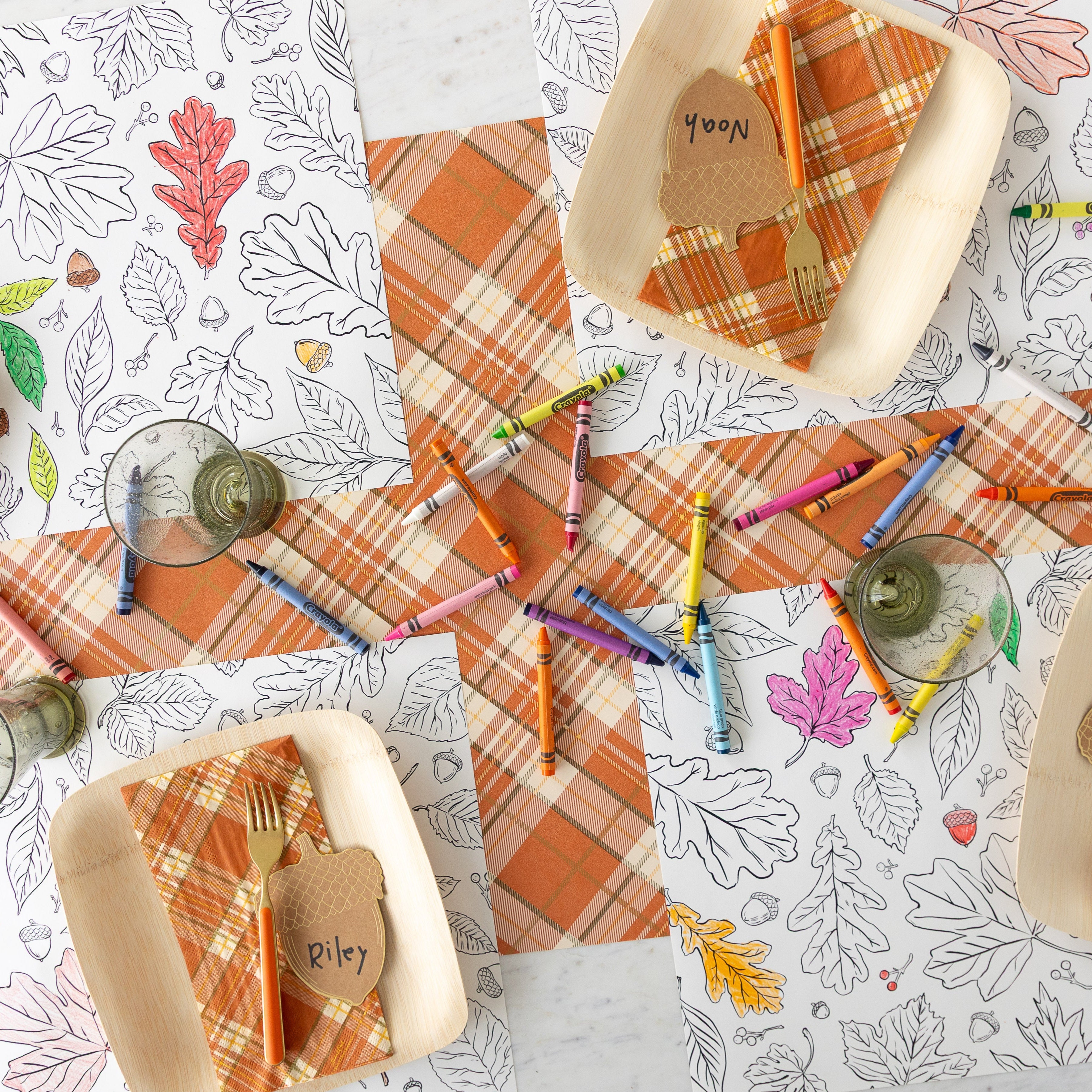 A place setting for four featuring the Coloring Leaves Placemats with a paper plate, fork and Acorn Place Card on top, scattered crayons on the table and Orange Plaid Runner underneath.