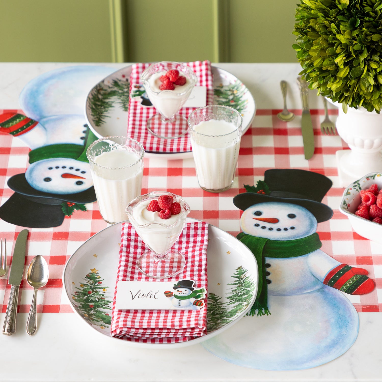 A festive table setting with the Die-cut Jolly Snowman Placemat, a festive tree plate, red checkered napkins with the Jolly Snowman place card and raspberry parfait on top and Red Painted Check Runner underneath.