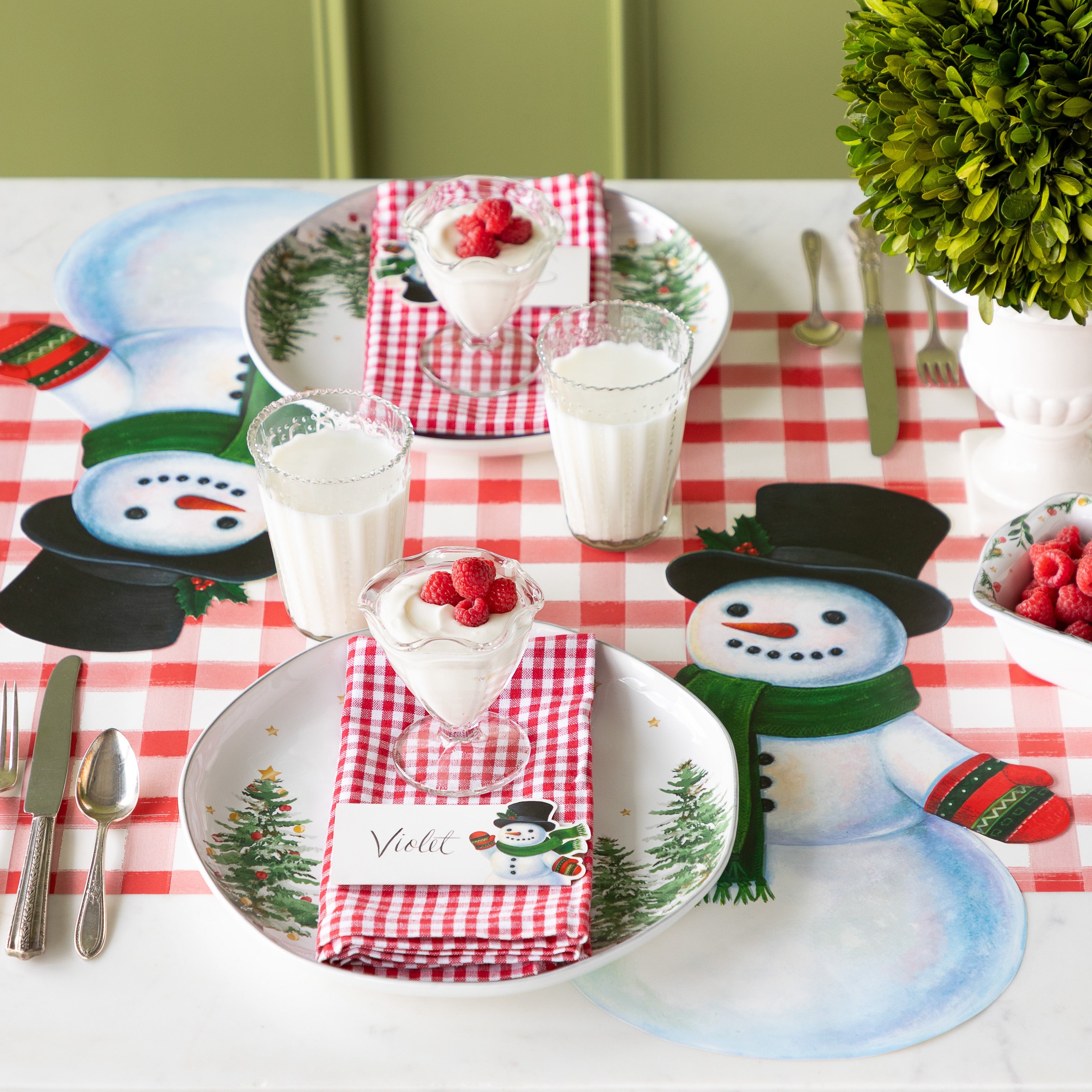 A festive table setting with the Die-cut Jolly Snowman Placemat, a festive tree plate, red checkered napkins with the Jolly Snowman place card and raspberry parfait on top and Red Painted Check Runner underneath.
