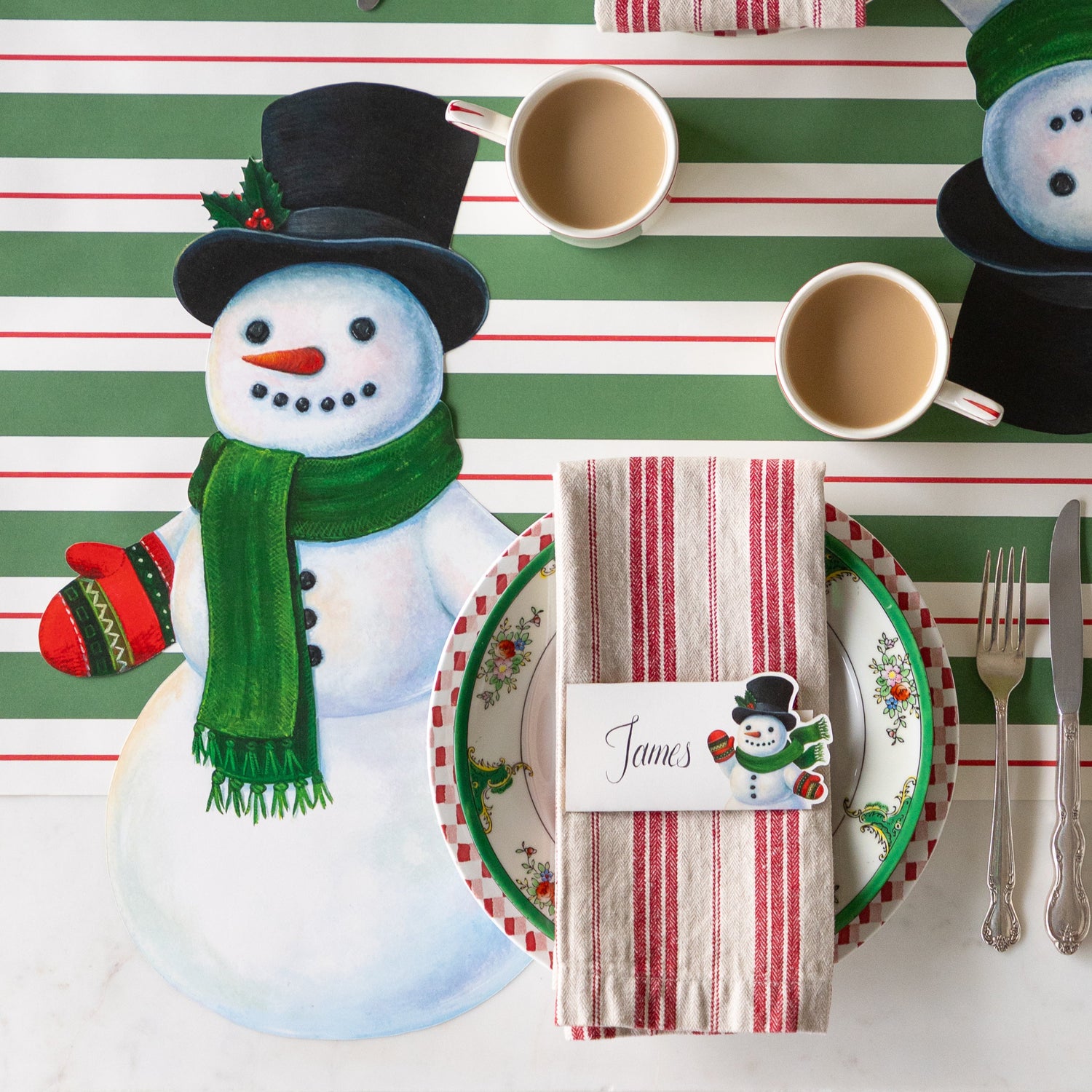 A festive place setting with the Die-cut Jolly Snowman Placemat, cup of coffee, red striped napkins with the Jolly Snowman Place Card on top and Green &amp; Red Awning Stripe Runner underneath.