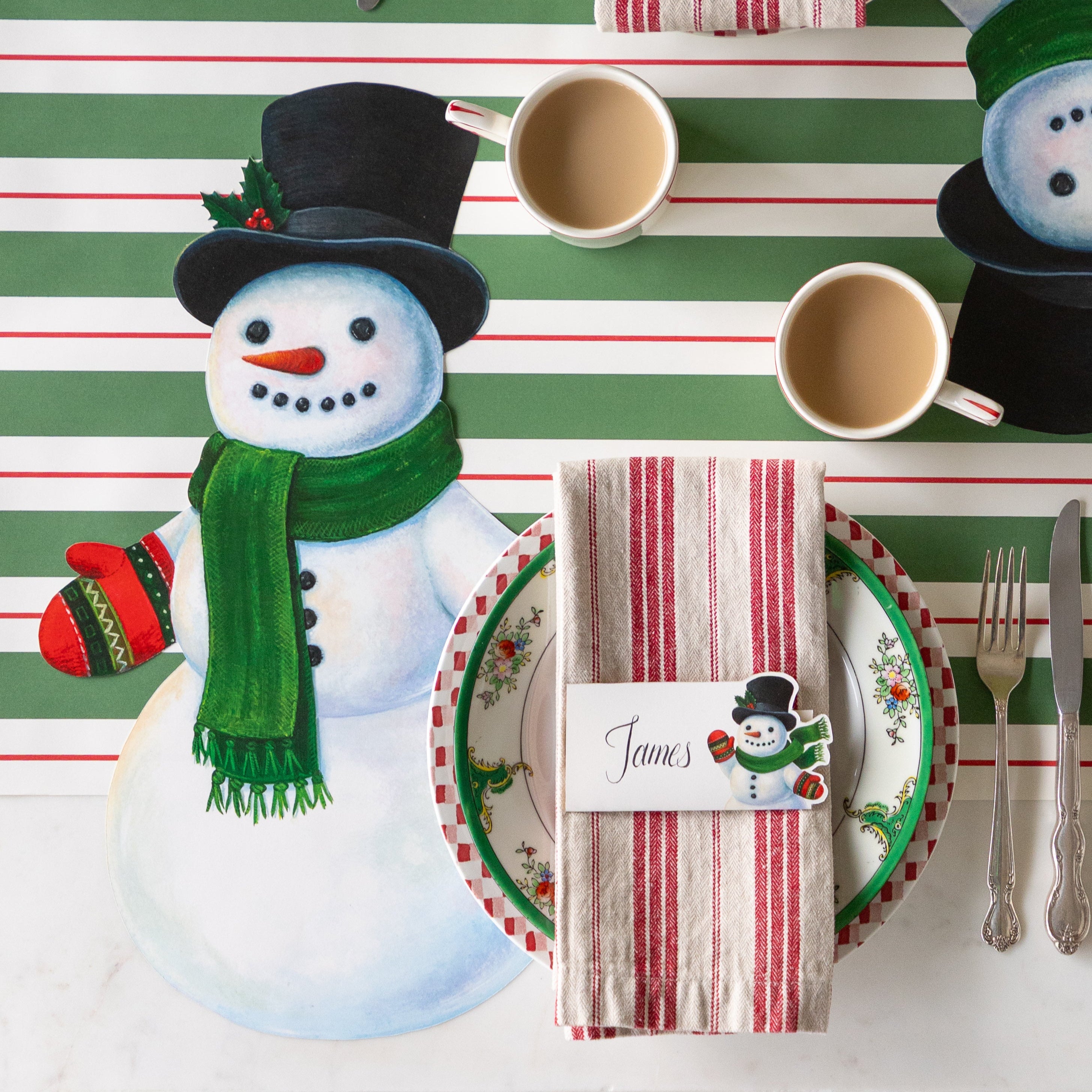 A festive place setting with the Die-cut Jolly Snowman Placemat, cup of coffee, red striped napkins with the Jolly Snowman Place Card on top and Green &amp; Red Awning Stripe Runner underneath.