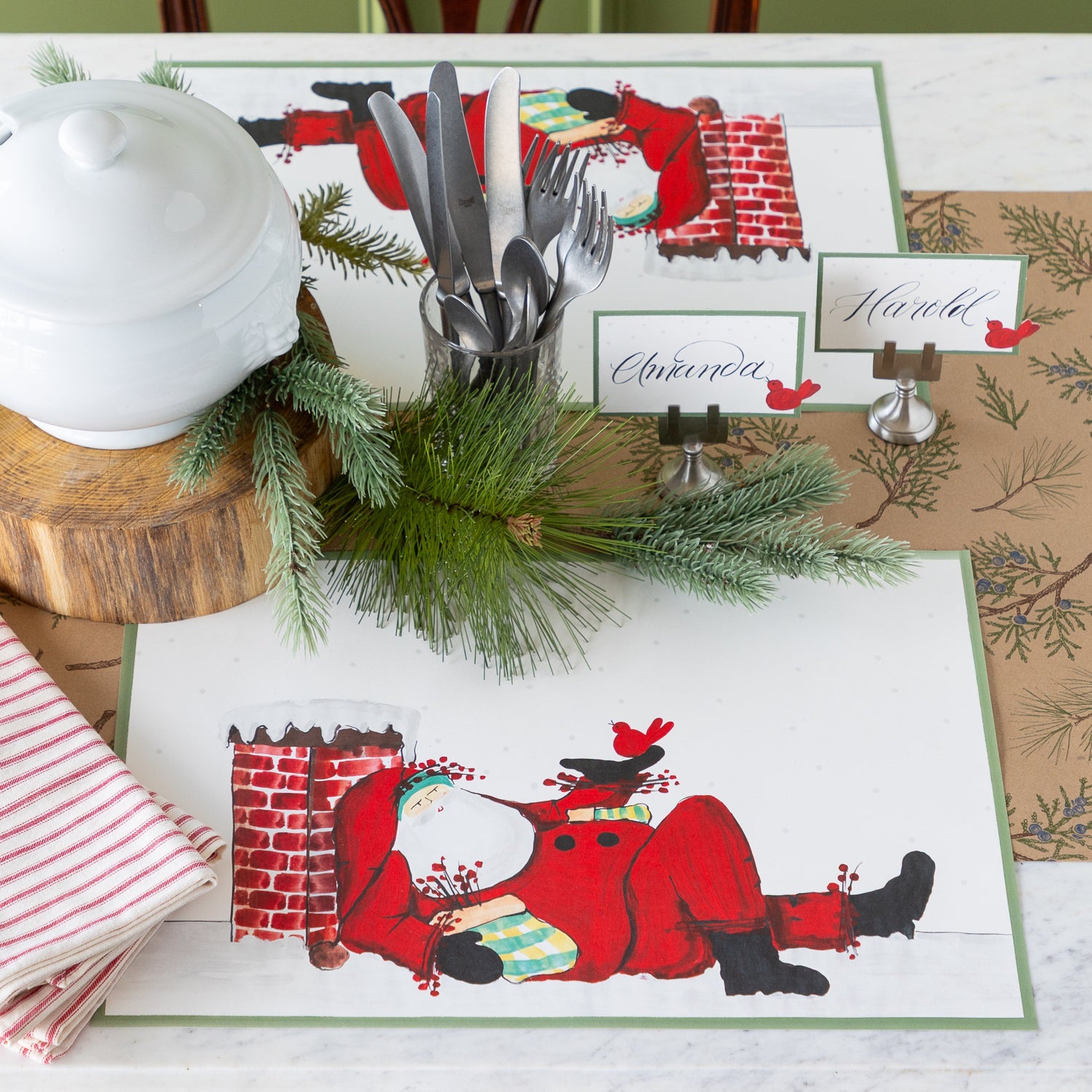 Christmas themed table setting featuring the Old St. Nick Chimney Placemats, red-and-white striped napkins, vintage silverware, Red Cardinal Place Cards and woodland decor on the Juniper Sprigs on Kraft Runner.