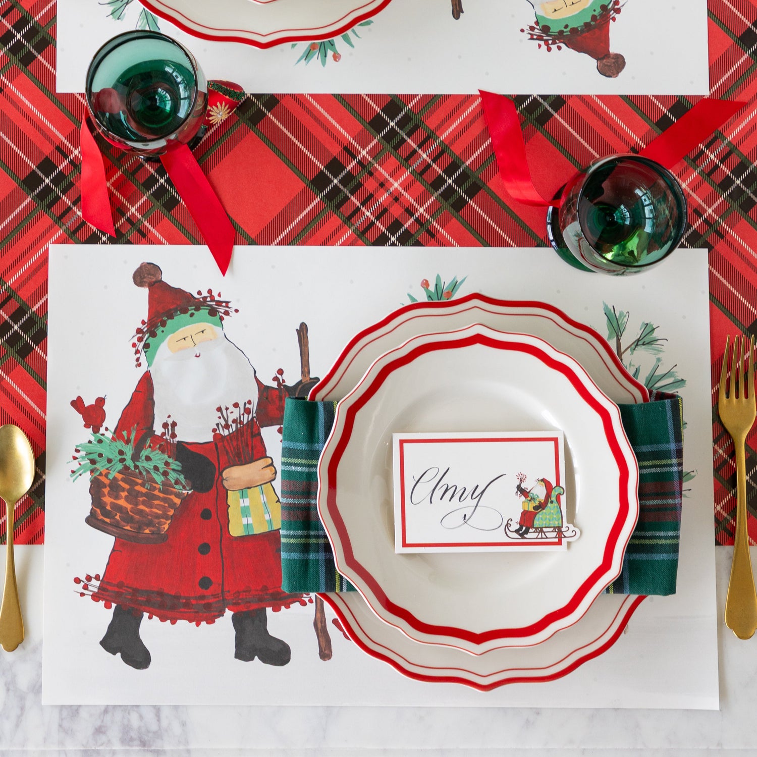 Christmas-themed table setting with Old St. Nick Forest Placemat, Red Icing on the Cake plates with the Old St. Nick Sleigh Place Card atop, on the Red Plaid Runner.