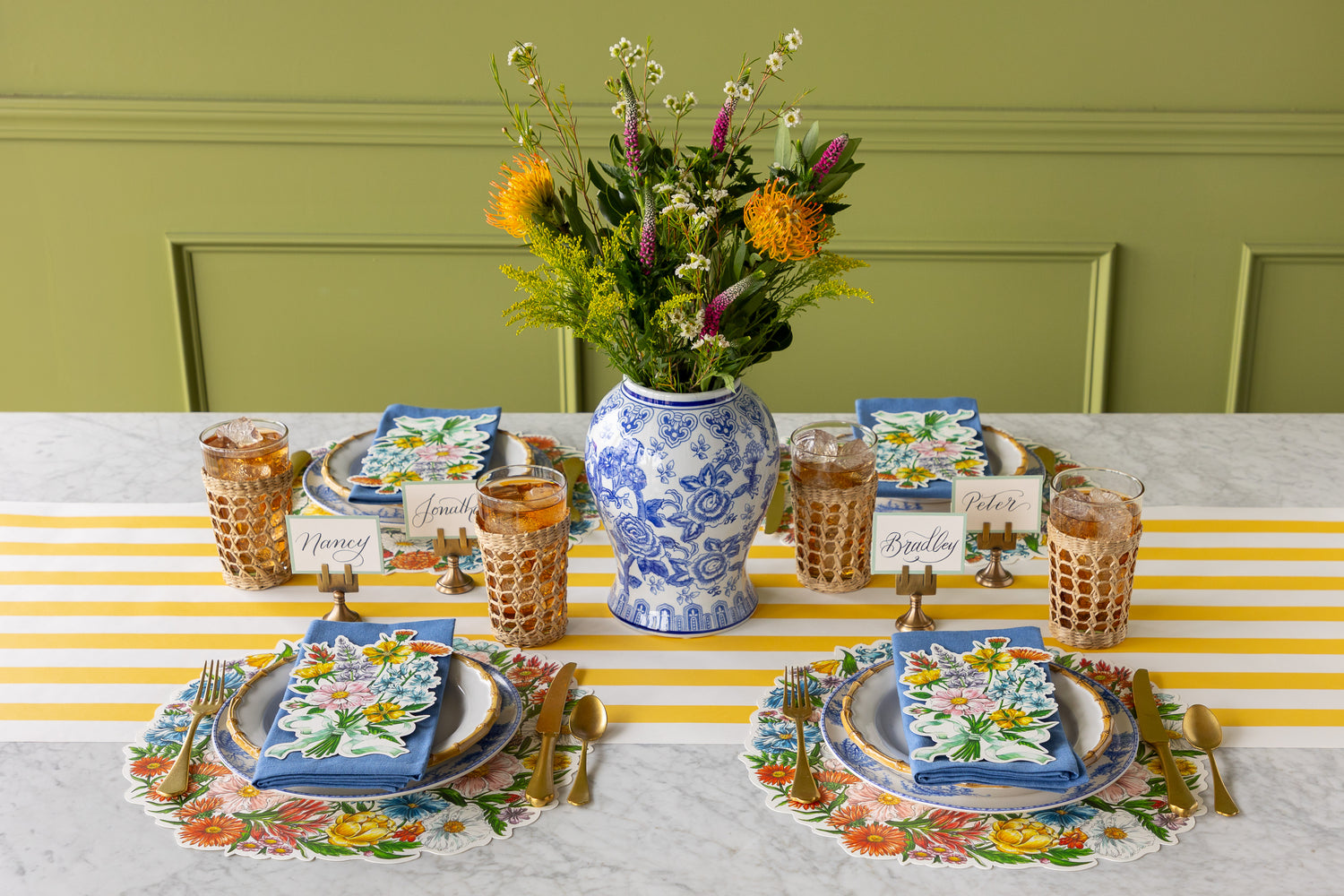 Table setting with Die-cut Wildflower Wreath Placemats, a floral arrangement in a vase, floral and bamboo plates, blue napkins, Wildflower Table Accents and gold cutlery on the Marigold Classic Stripe Runner.
