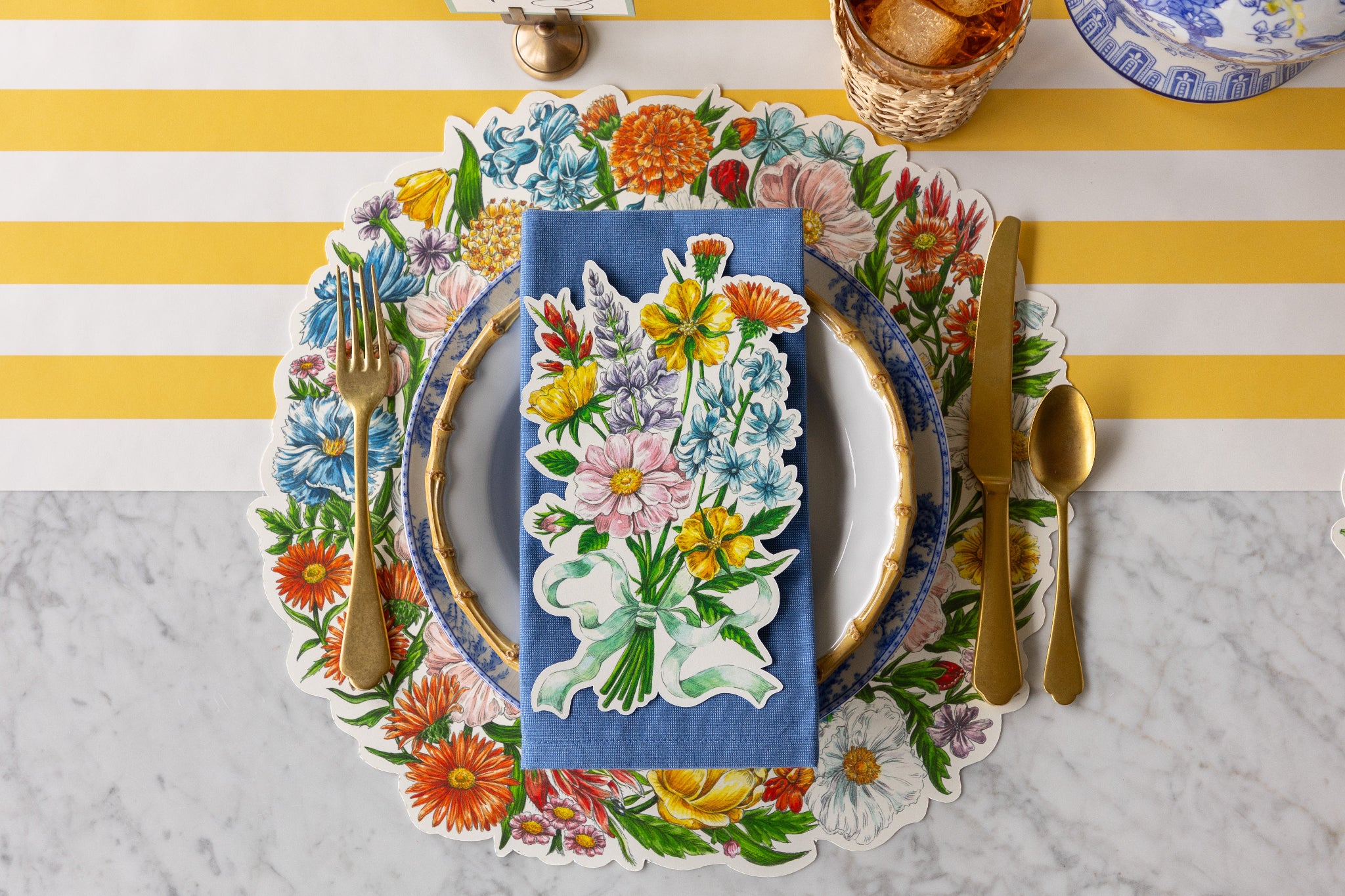 Table setting with Die-cut Wildflower Wreath Placemat, floral and bamboo plates, blue napkin, Wildflower Table Accent and gold cutlery on the Marigold Classic Stripe Runner.