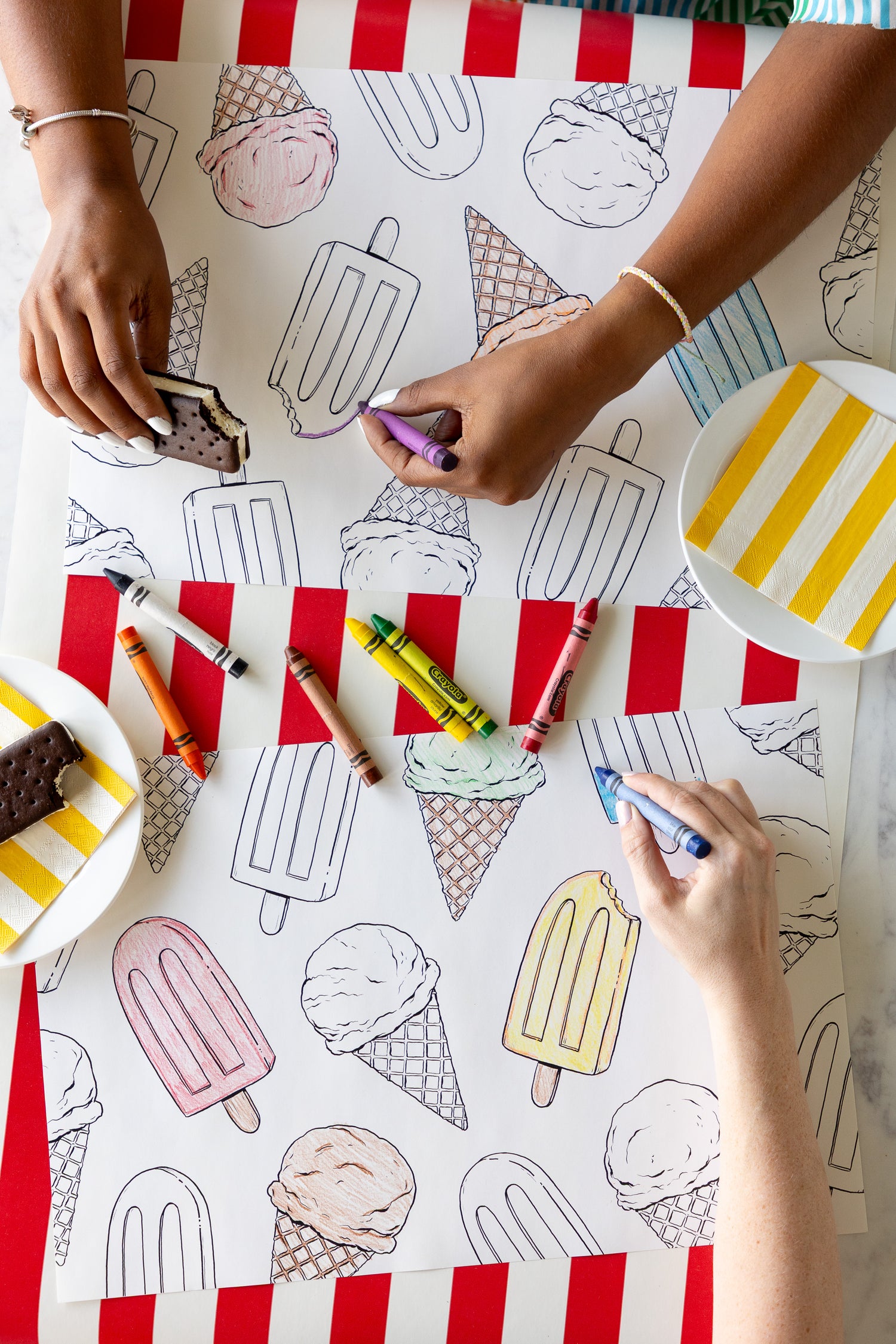 Two people coloring the Coloring Summer Treats Placemats with crayons on a table with the Red Classic Stripe Runner.