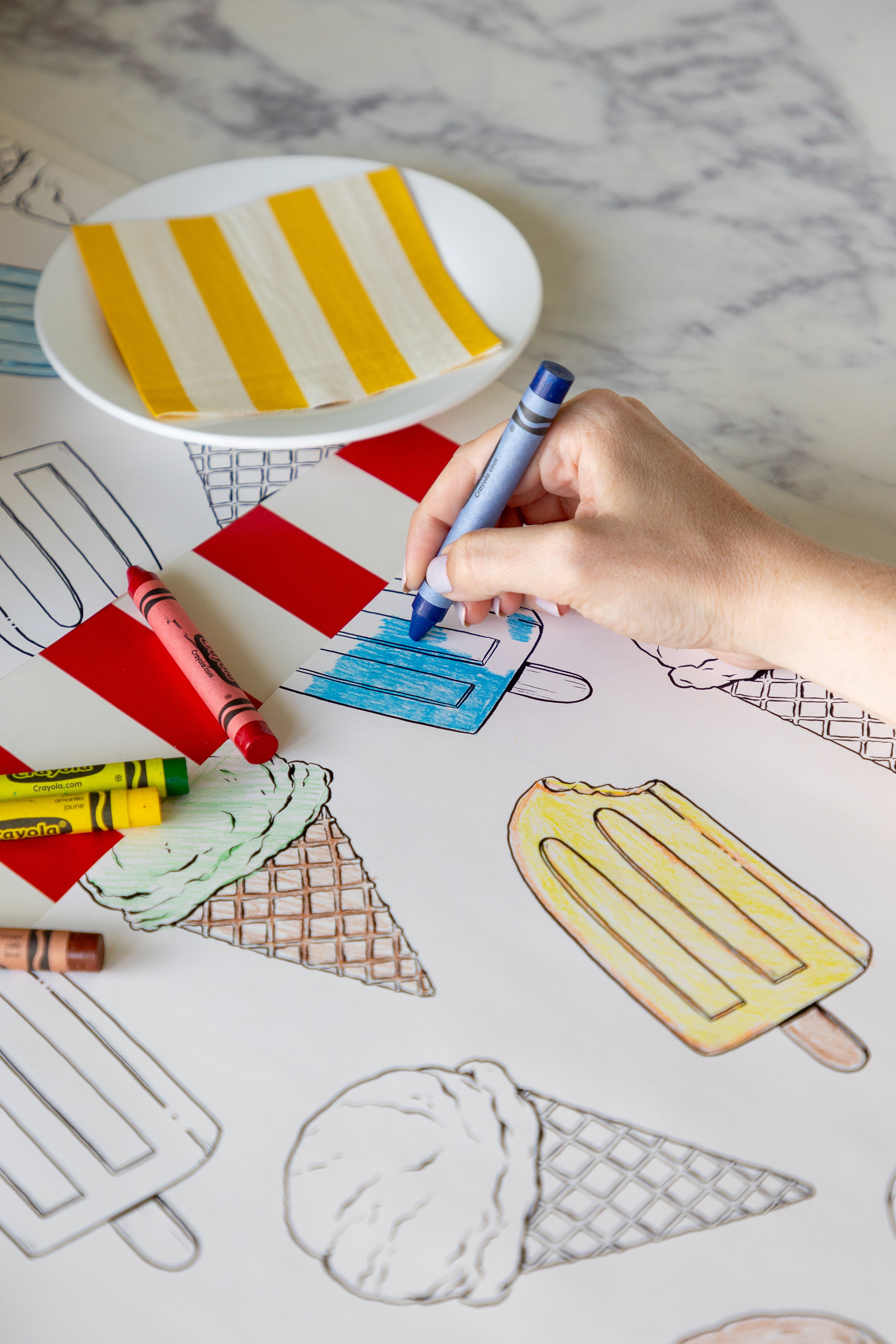 A person coloring the Coloring Summer Treats Placemat with crayons on a table with the Red Classic Stripe Runner.