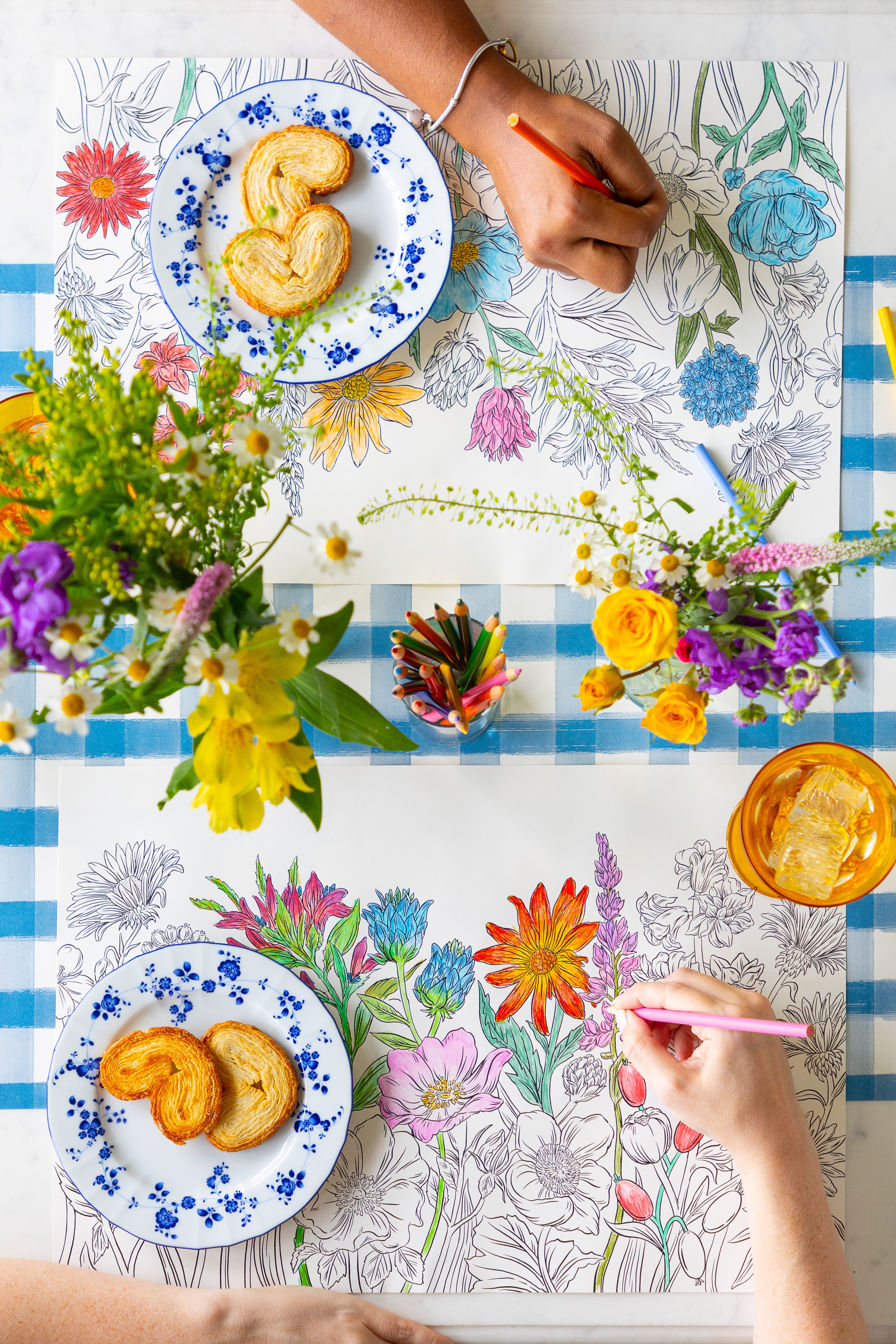 Two people coloring the Coloring Wildflower Meadow Placemat on a table with plates of food and flowers.