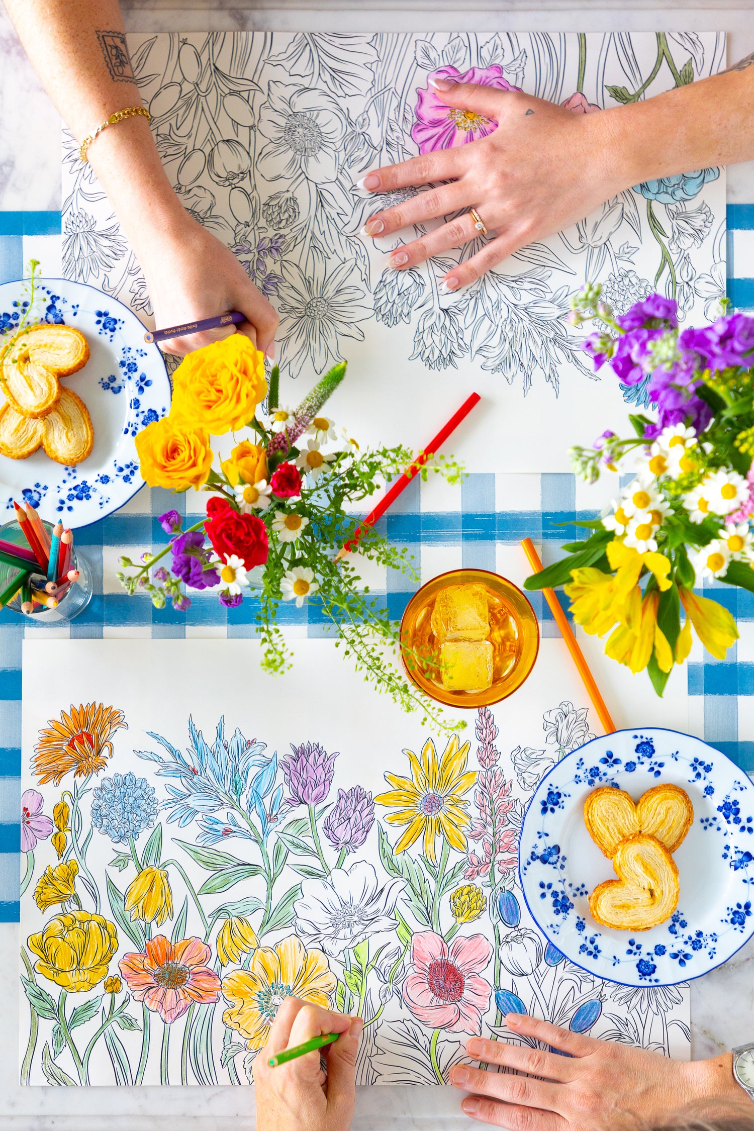 Two people coloring the Coloring Wildflower Meadow Placemat on a table with plates of food and flowers.