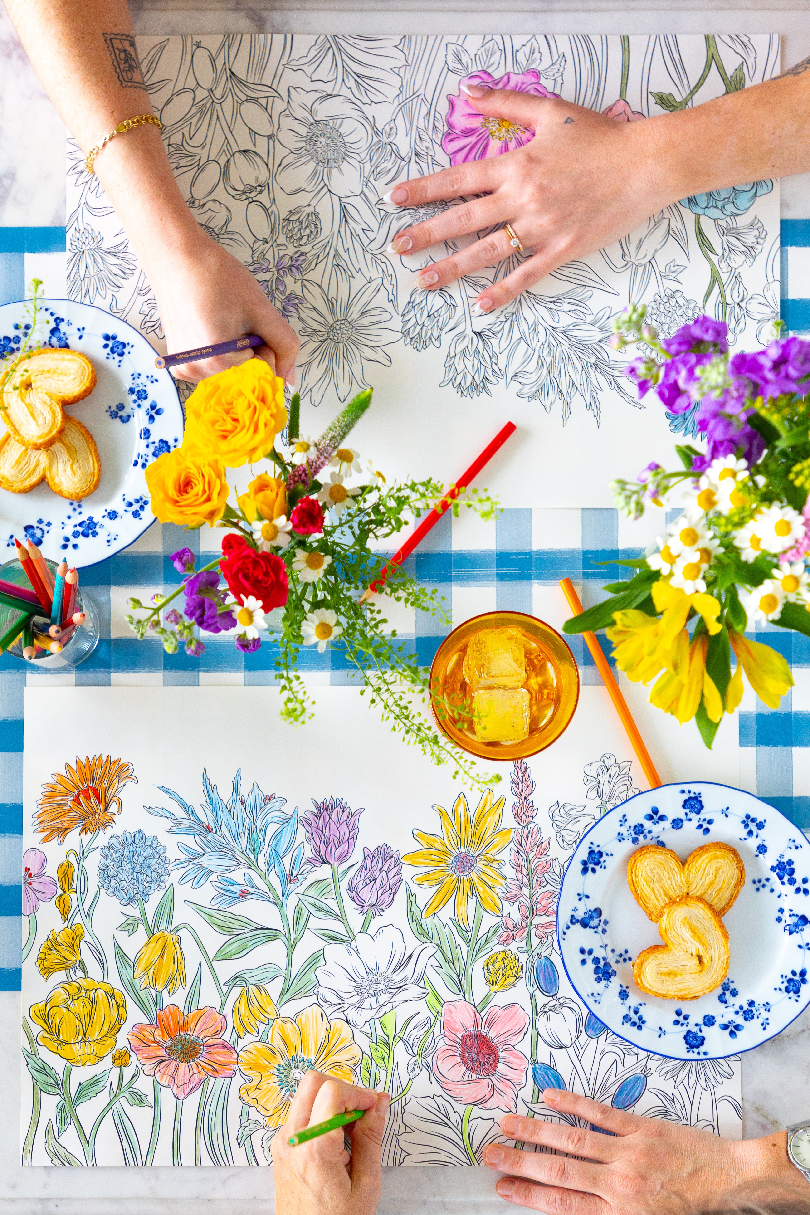 Two people coloring the Coloring Wildflower Meadow Placemat on a table with plates of food and flowers.