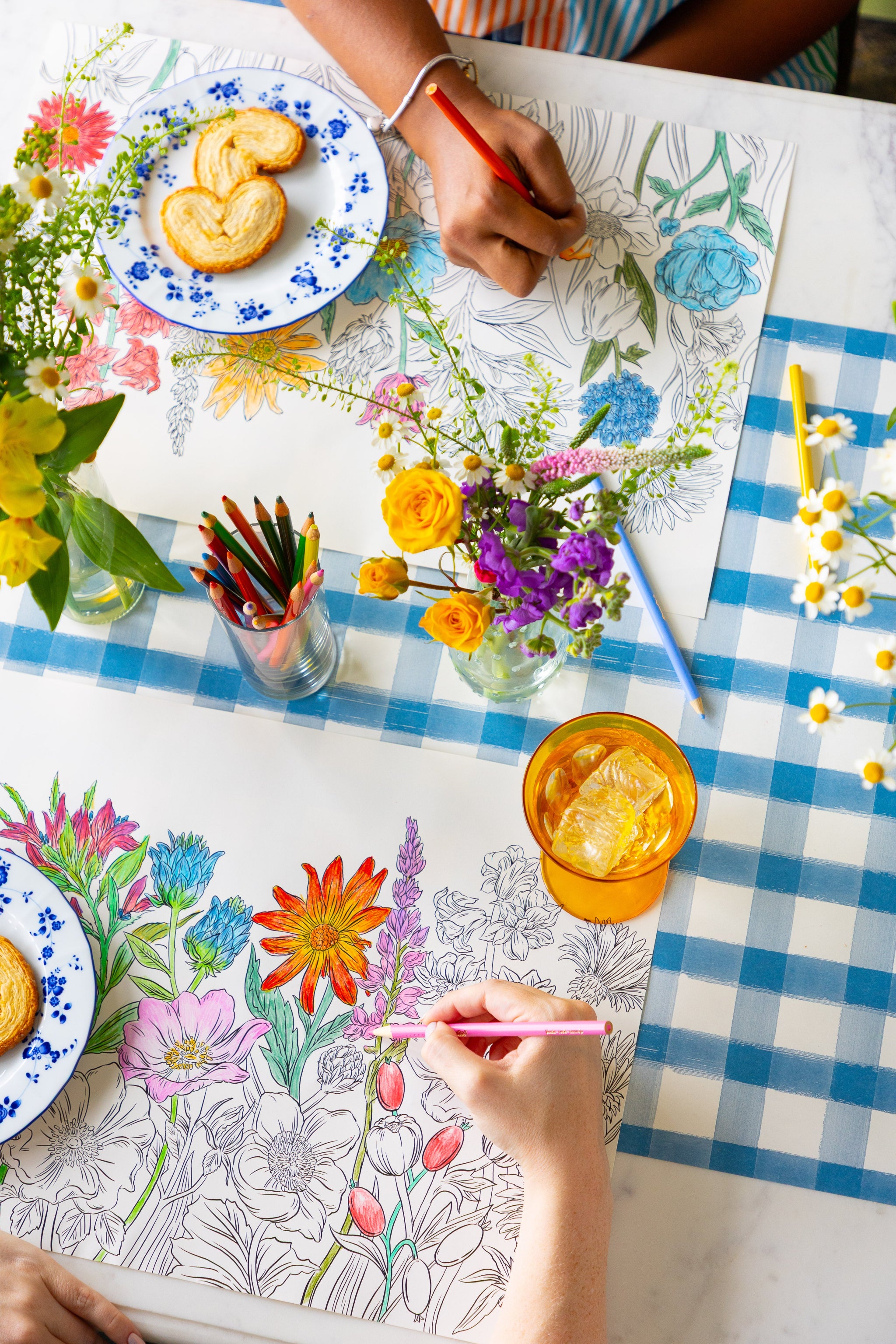 Two people coloring the Coloring Wildflower Meadow Placemat on a table with plates of food and flowers.