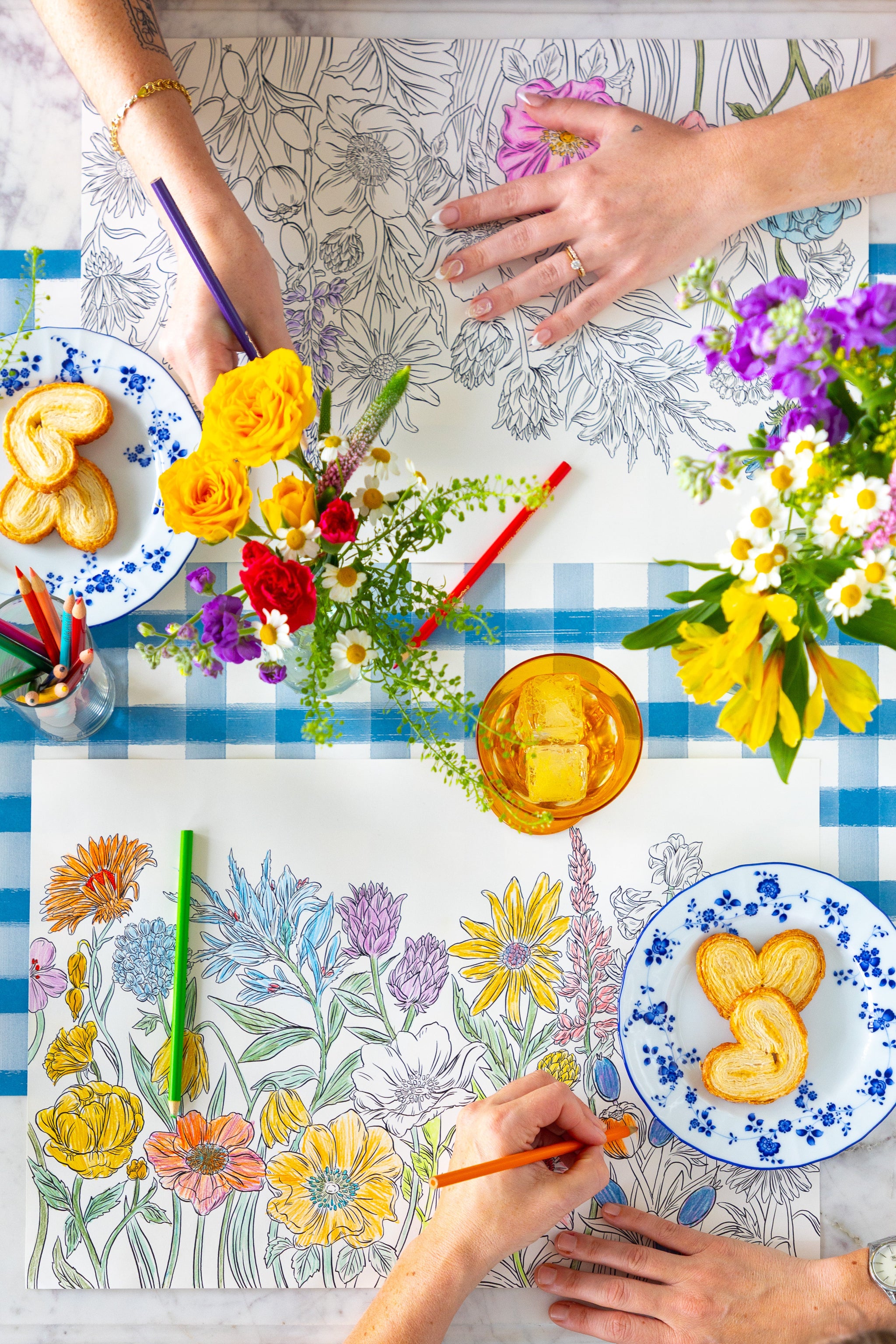Two people coloring the Coloring Wildflower Meadow Placemat on a table with plates of food and flowers.