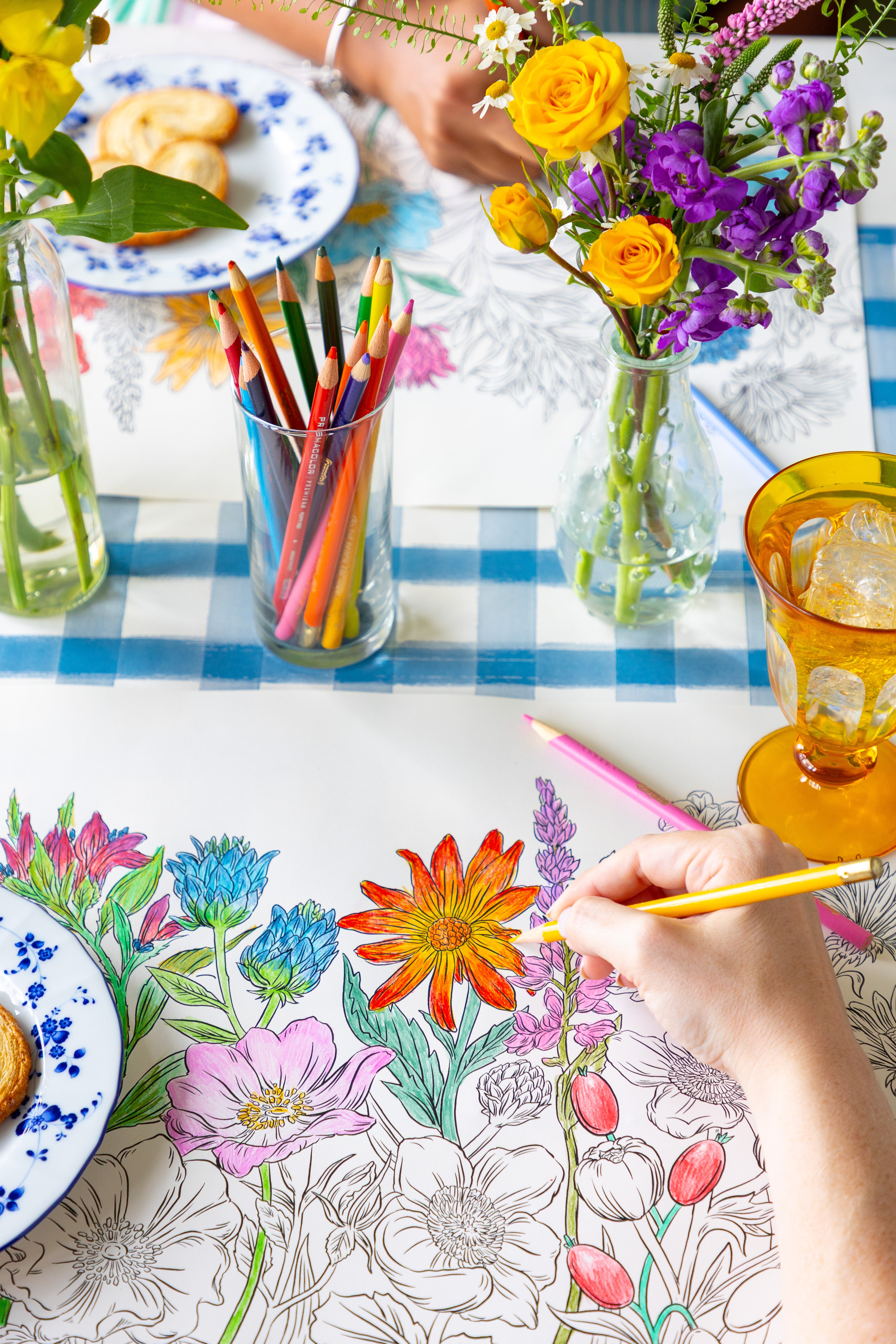 Two people coloring the Coloring Wildflower Meadow Placemat on a table with plates of food and flowers.