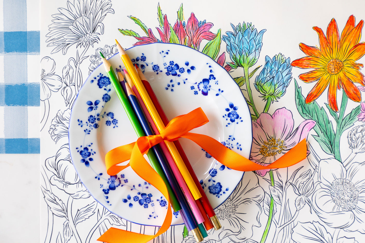 Coloring Wildflower Meadow Placemat partially colored in with a floral plate and colored pencils tied together by a ribbon atop.