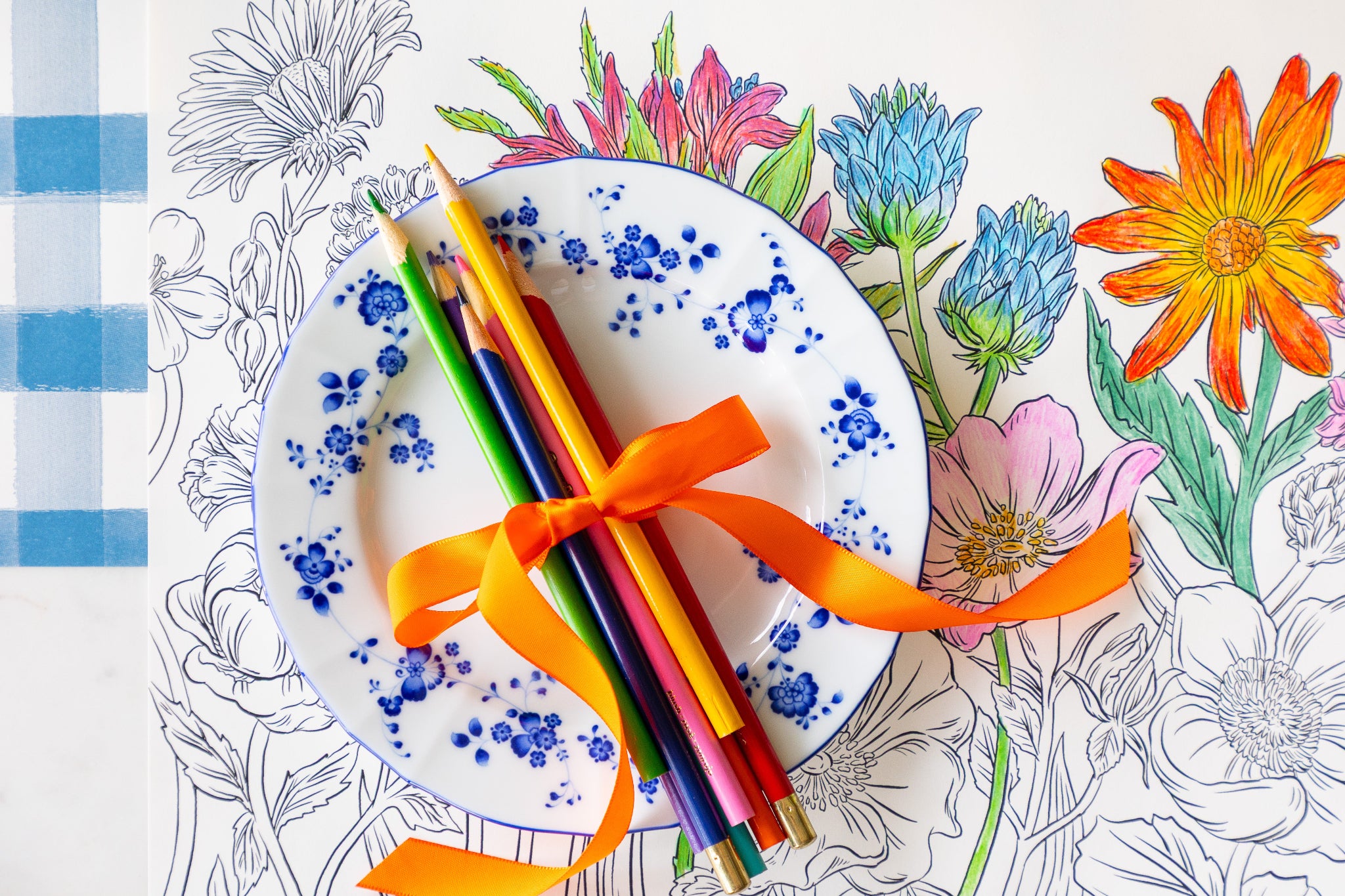 Coloring Wildflower Meadow Placemat partially colored in with a floral plate and colored pencils tied together by a ribbon atop.