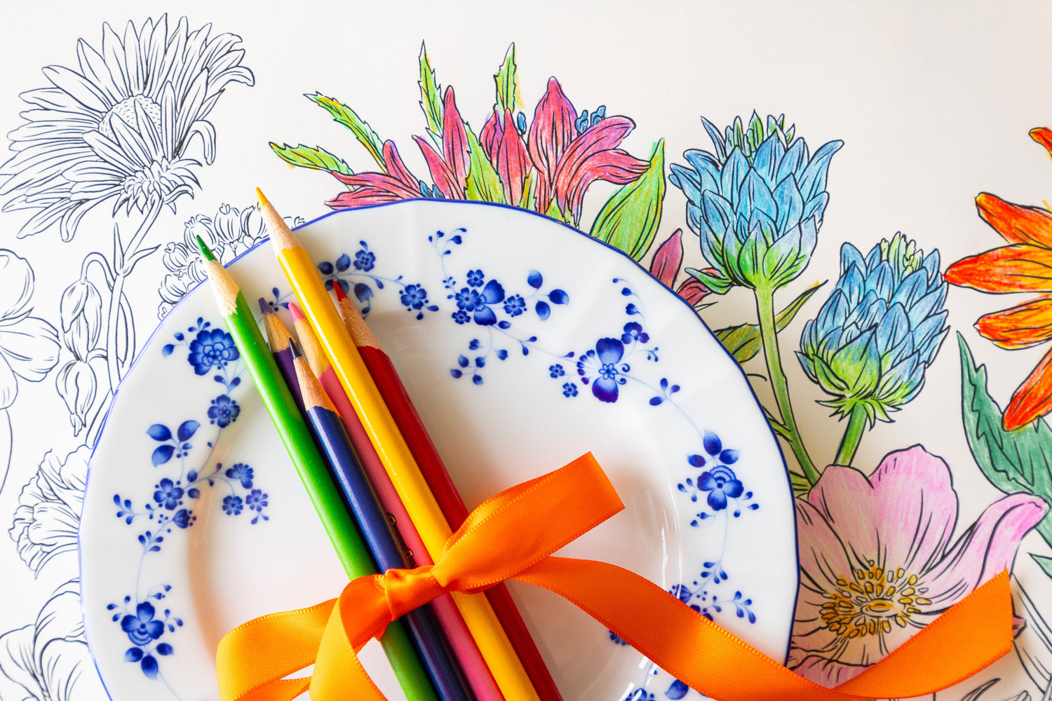 Coloring Wildflower Meadow Placemat partially colored in, with colored pencils and a plate on the Blue Painted Check Runner.