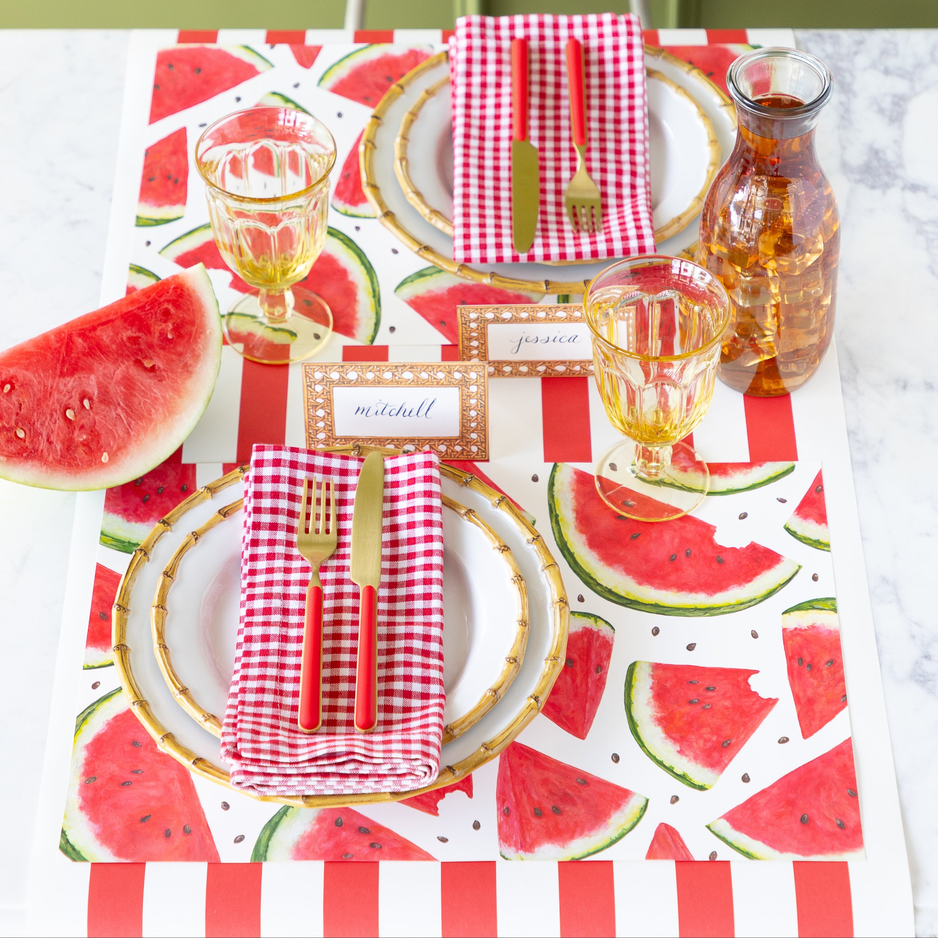Dining table setting with  Watermelon Picnic Placemats, bamboo rimmed plates, red checkered napkins, Rattan Weave Place Cards and red and gold flatware on the Red Classic Stripe Runner.