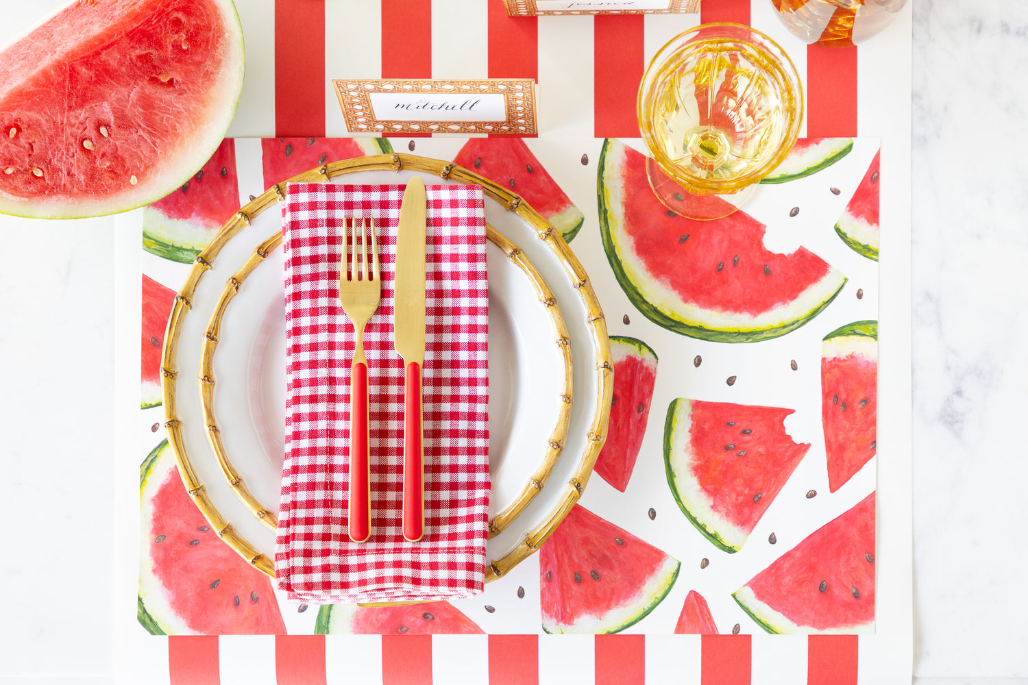 Dining table setting with Watermelon Picnic Placemat, bamboo rimmed plates, red checkered napkin, Rattan Weave Place Card and red and gold flatware on the Red Classic Stripe Runner.