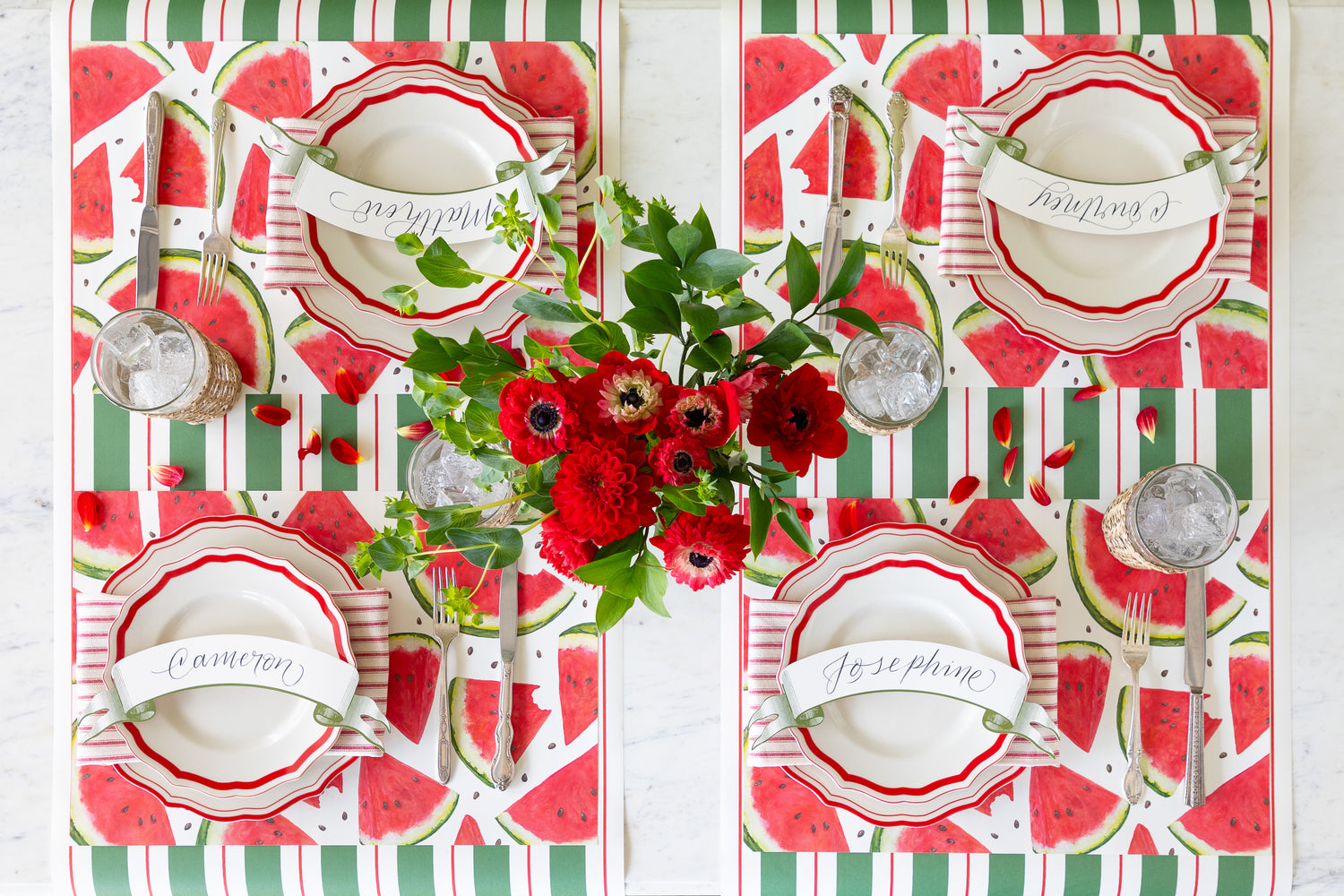 Decorative table setting with red and green watermelon-themed placemats, white plates, and floral centerpieces.