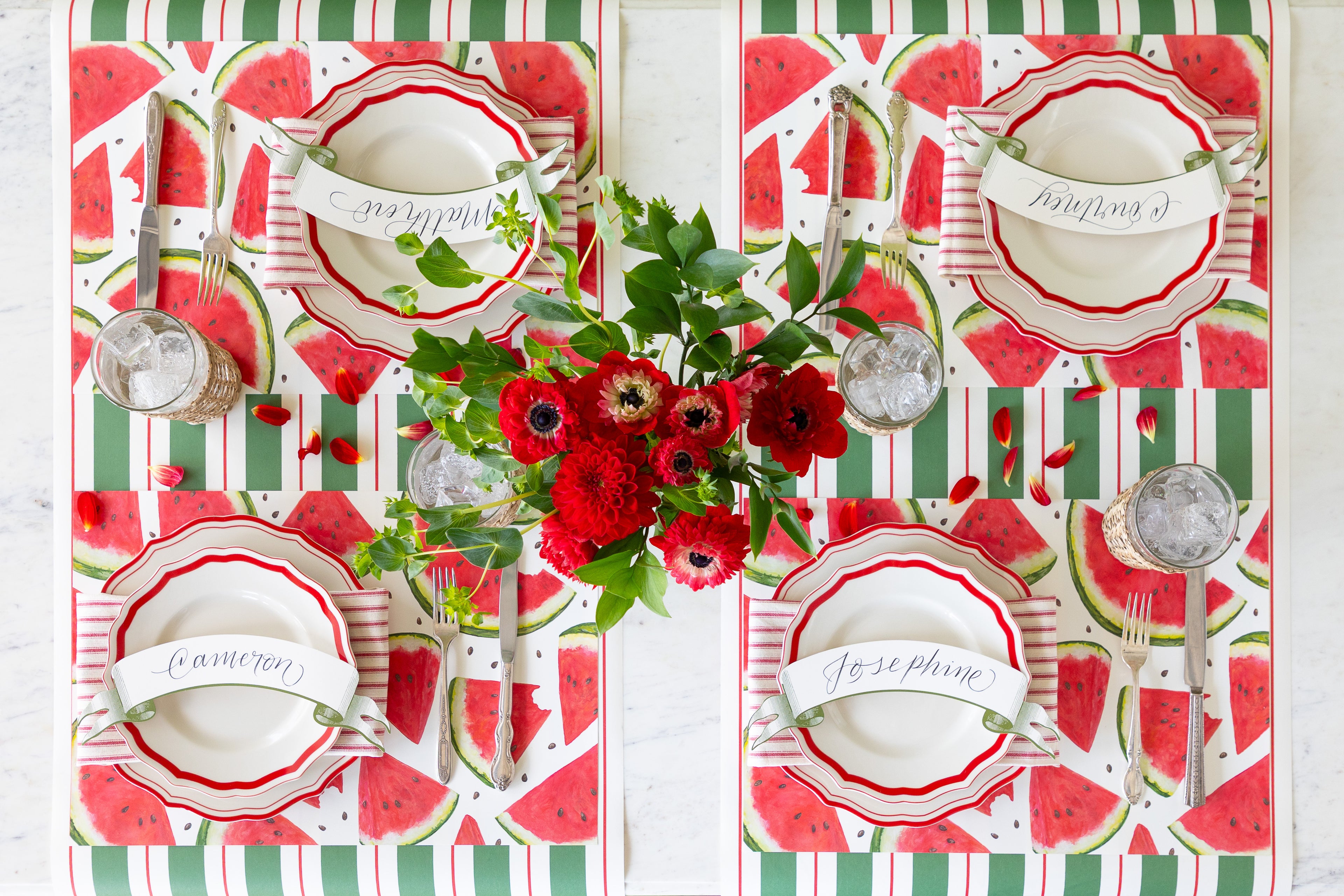 Decorative table setting with red and green watermelon-themed placemats, white plates, and floral centerpieces.