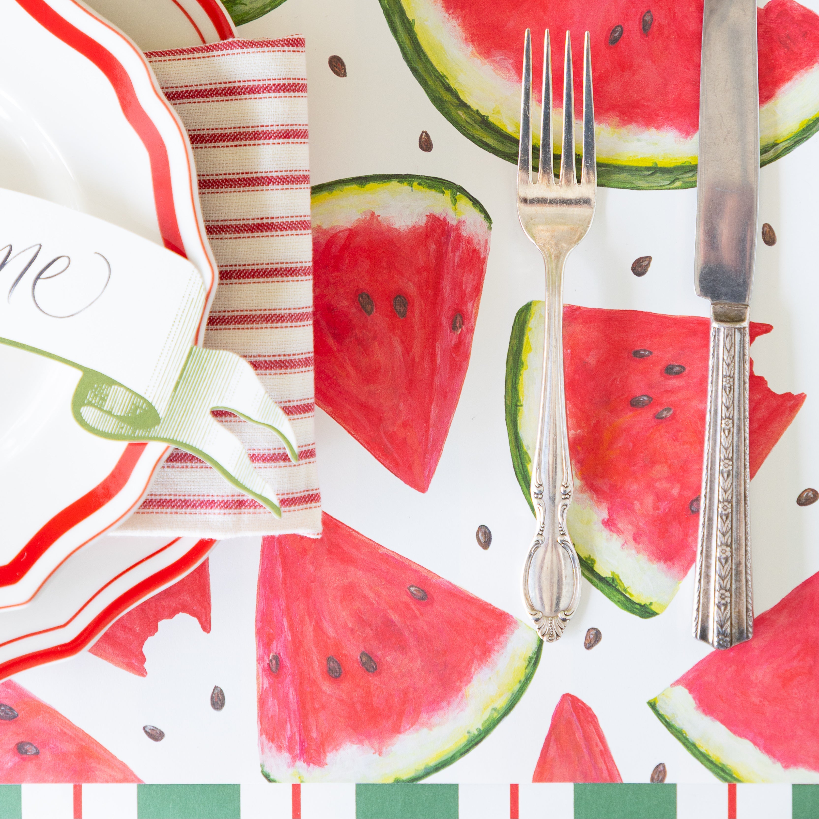 Table setting with watermelon-themed placemats, cutlery, and a glass of water.