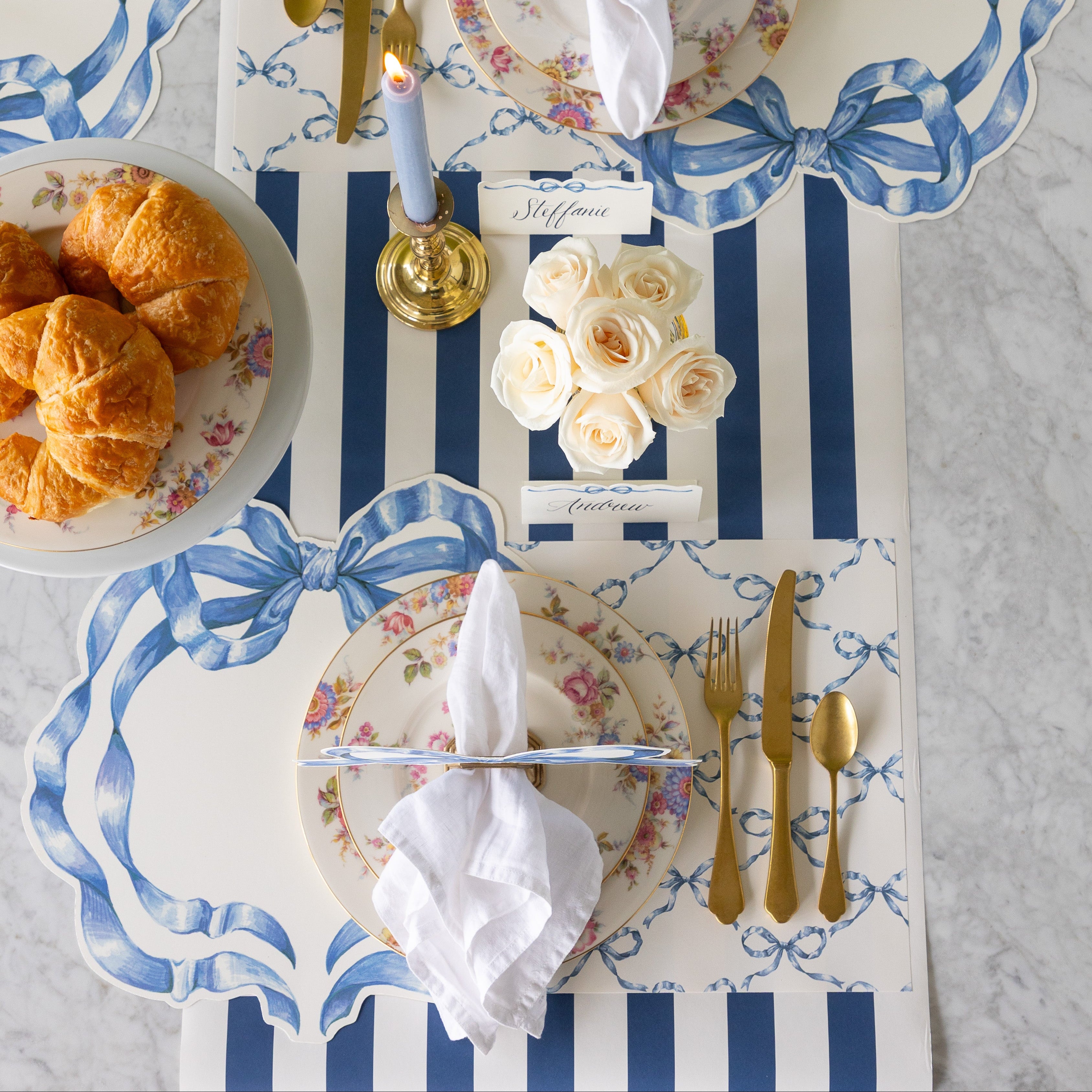 Elegant table setting with flowers, a candle and croissants, Die-cut Heirloom Blue Bow and Heirloom Blue Bow Lattice Placemats with coordinating blue bow items.