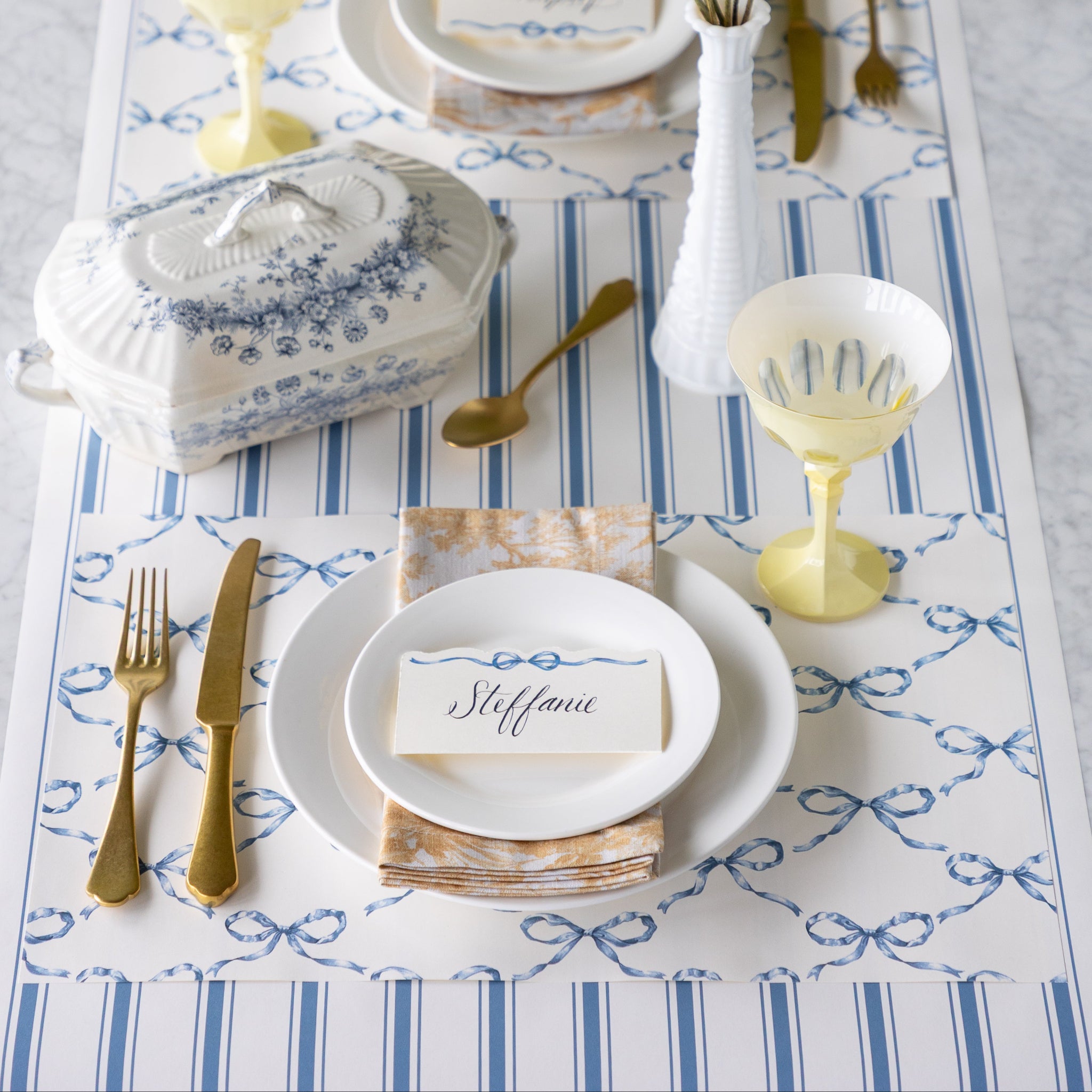 Elegant table setting with Heirloom Blue Bow Lattice Placemat, Heirloom Blue Bow Place Card, Cornflower Blue Linen Stripe Runner, white plates, gold cutlery, and decorative elements.
