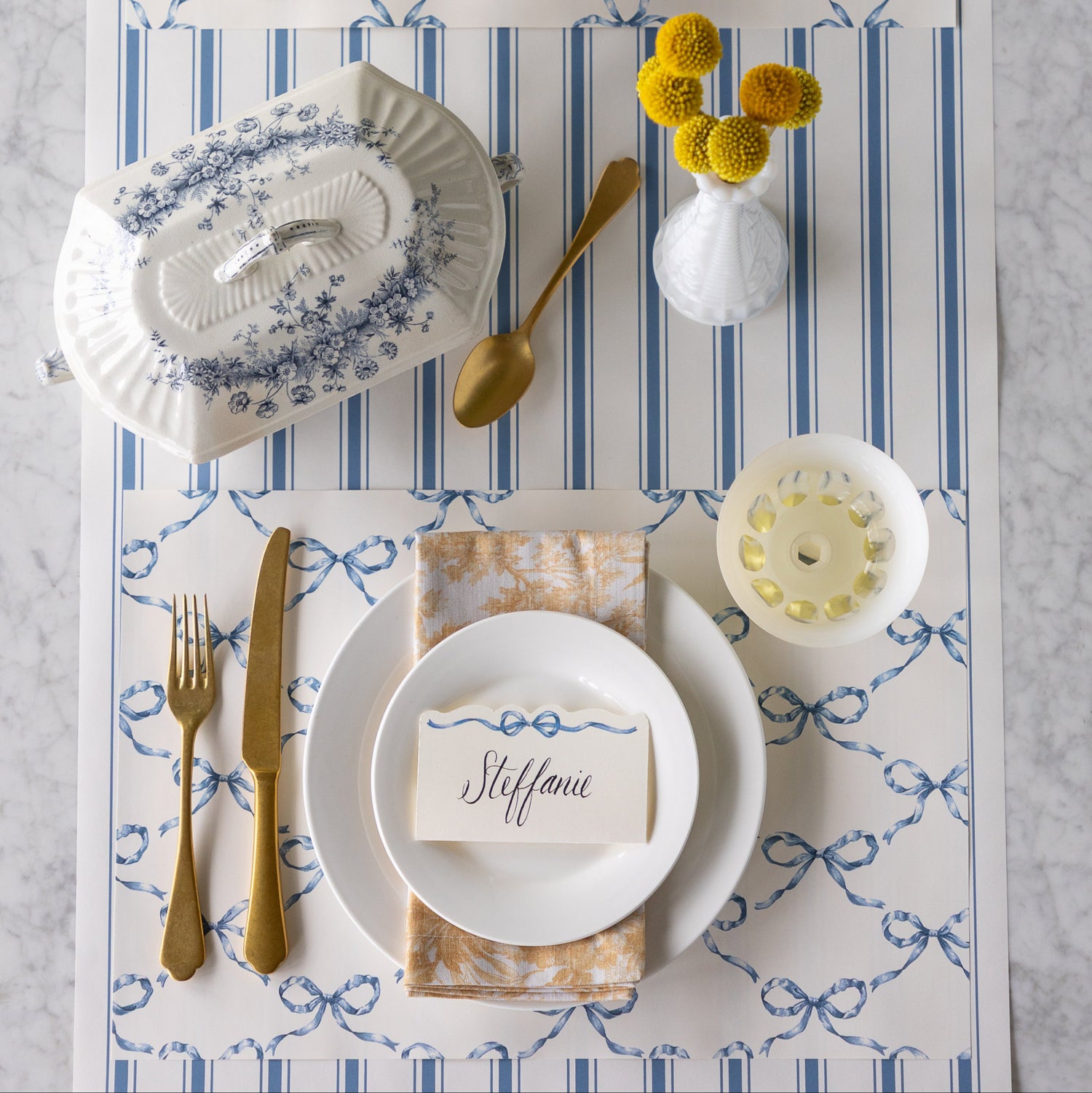 Elegant table setting with Heirloom Blue Bow Lattice Placemat, Heirloom Blue Bow Place Card, Cornflower Blue Linen Stripe Runner, white plates, gold cutlery, and decorative elements.