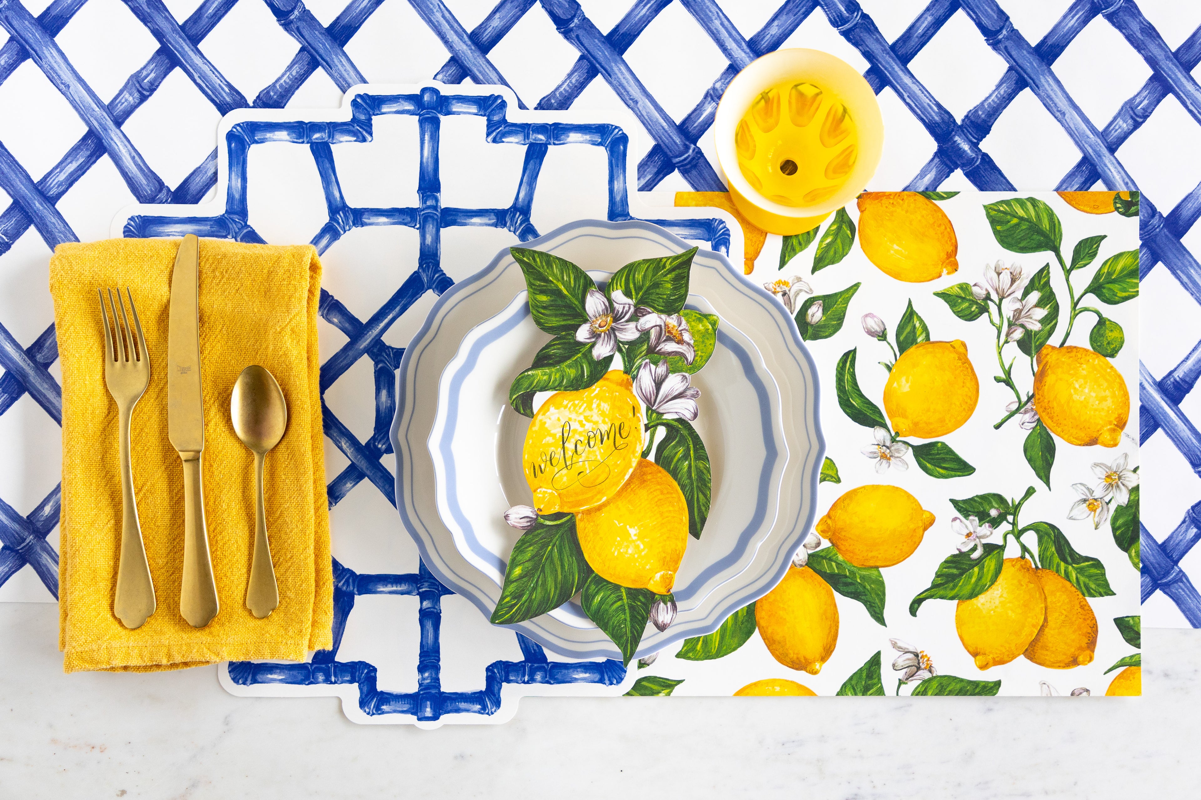 Table setting with the Die-cut Blue Bamboo and Lemons Placemats, Lemons Table Accent, Cornflower Blue Icing on the Cake plates, gold flatware and yellow napkin on the Blue Lattice Runner.