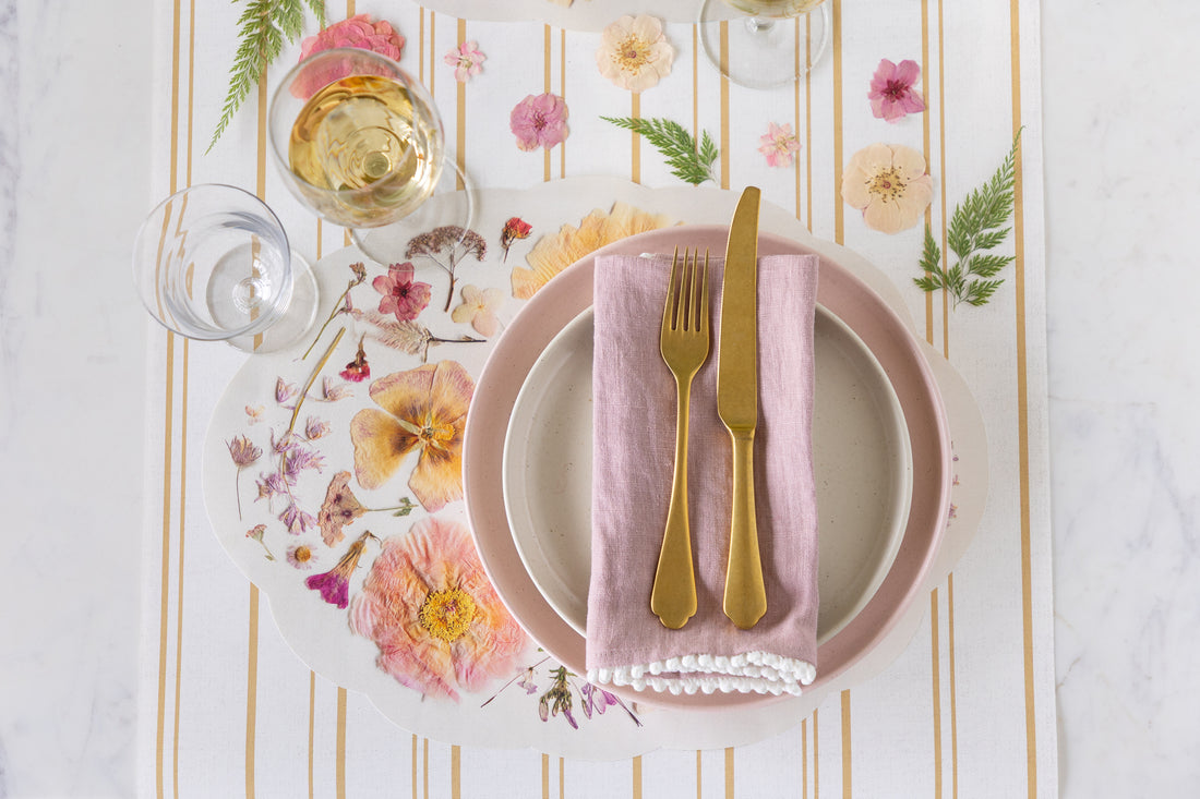 Dinner setting with Die-cut Blush Petals Placemat, pink plates, gold cutlery, and glasses on the Antique Gold Stripe Runner.