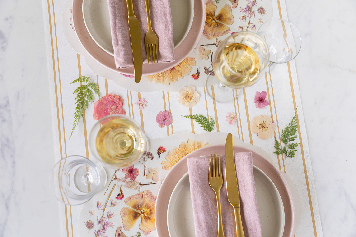 Table setting with floral placemats, pink plates, gold cutlery, and white wine glasses.