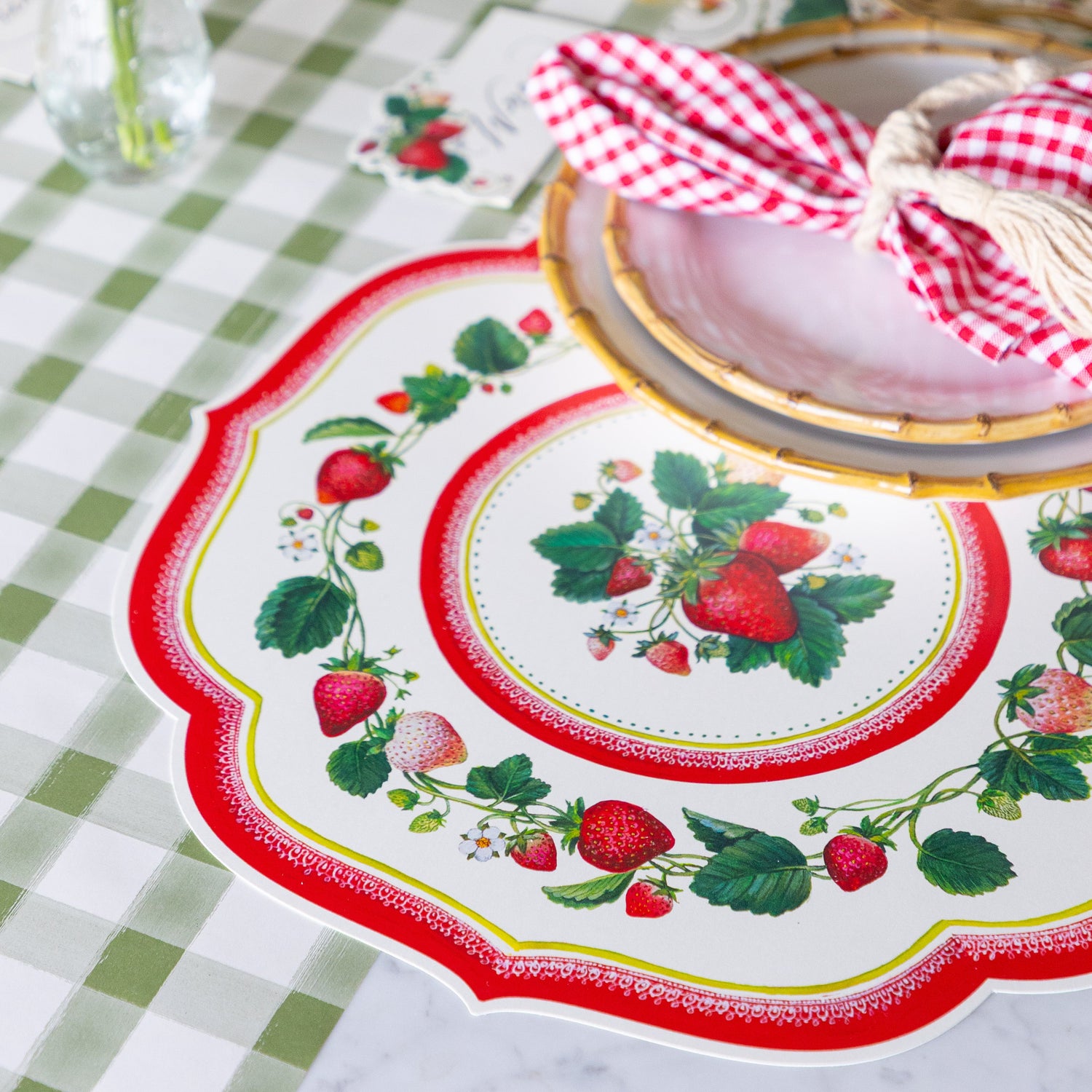 Decorative table setting with Die-cut Strawberry China Placemat, red checkered napkin, and gold cutlery on the Moss Painted Check Runner.