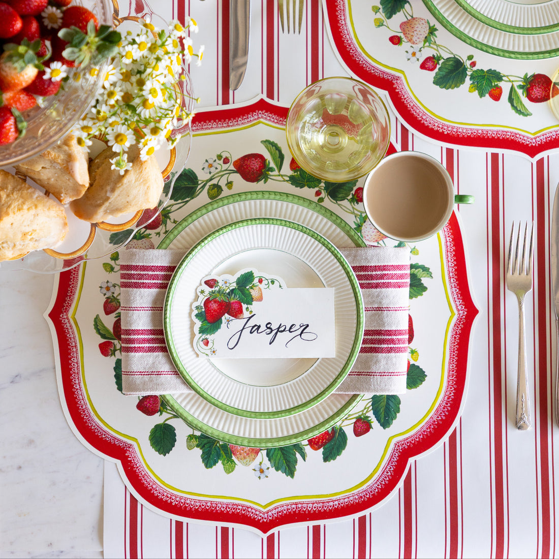 Decorative table setting with strawberry-themed plates and place card on a striped tablecloth.