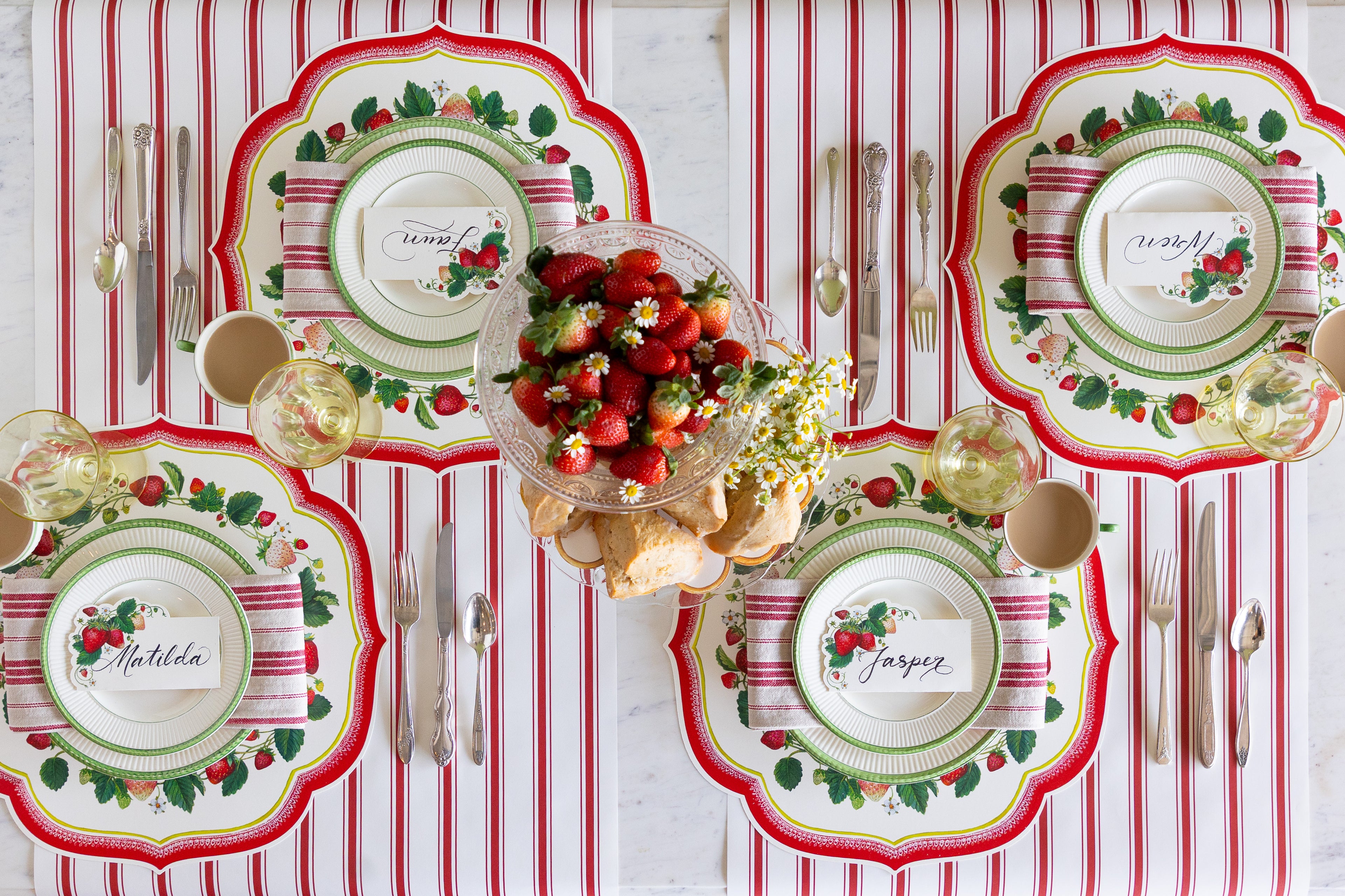 Decorative table setting with strawberries, flowers and scones, Die-cut Strawberry China Placemats, green rimmed plates, red striped napkins, Strawberries Place Cards and vintage cutlery on the Red Linen Stripe Runner.