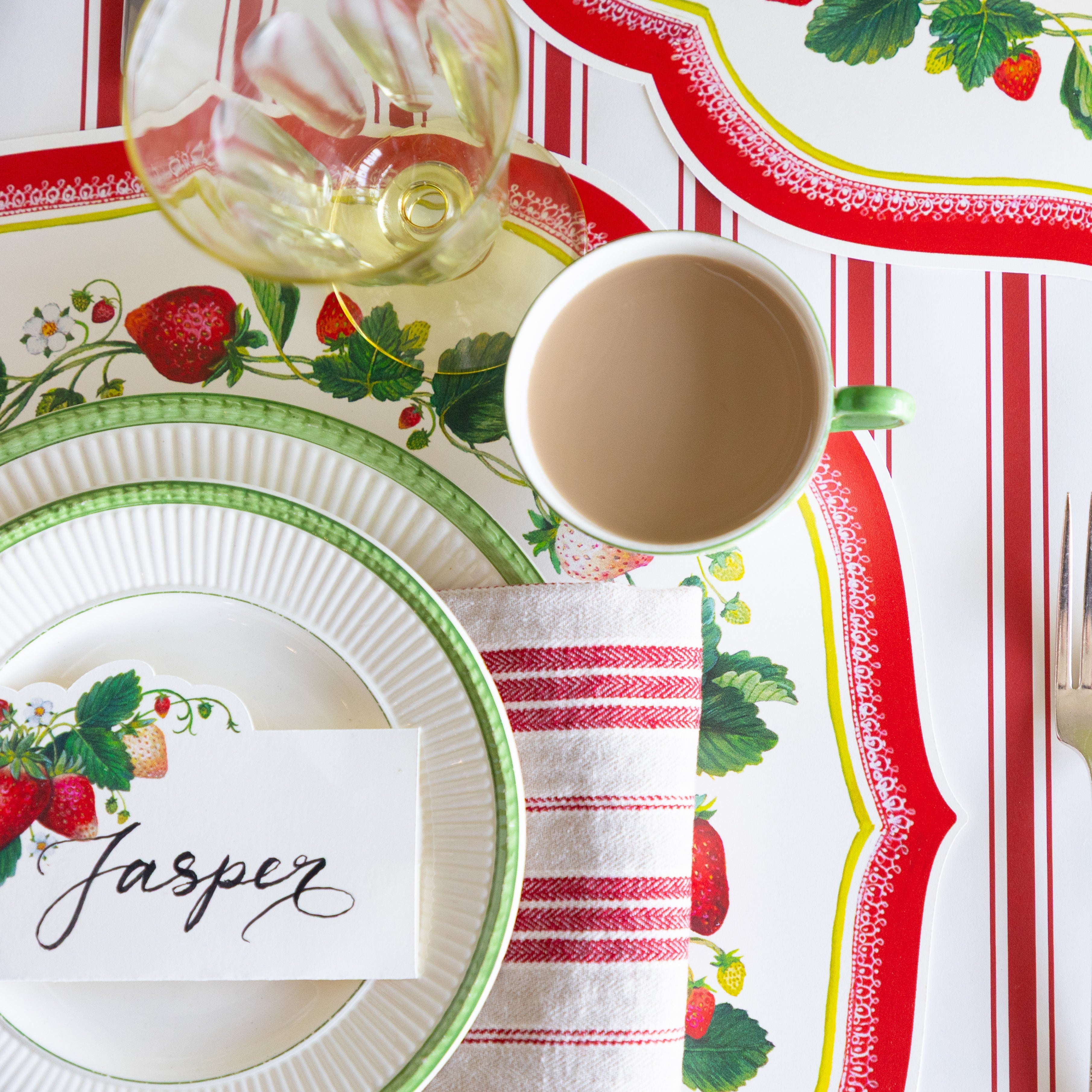 The Die-cut Strawberry China Placemat with green rimmed plates, Strawberries Place Card and red striped napkin on the Red Linen Stripe Runner.