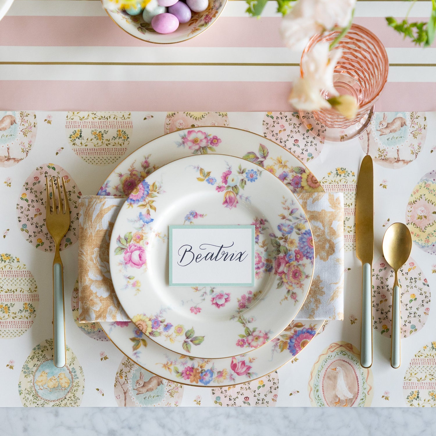 Easter table setting with floral plates, gold cutlery, Sweet Spring Eggs Placemat and Seafoam Frame Place Card on the Pink &amp; Gold Awning Stripe Runner.