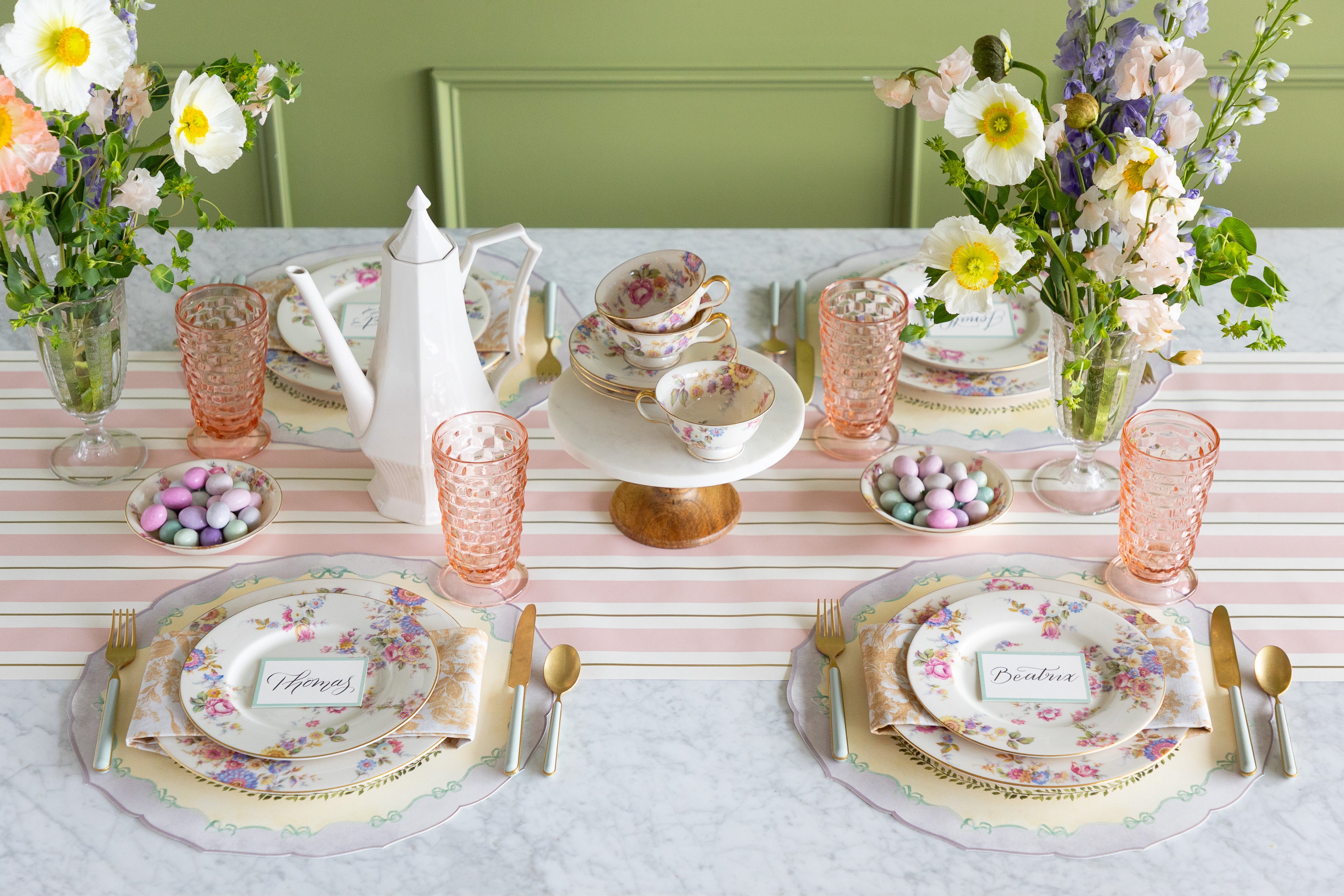 Easter table setting with flowers, floral plates, pastel glasses, Easter eggs and Die-cut Spring Meadow Placemats on the Pink &amp; Gold Awning Stripe Runner.