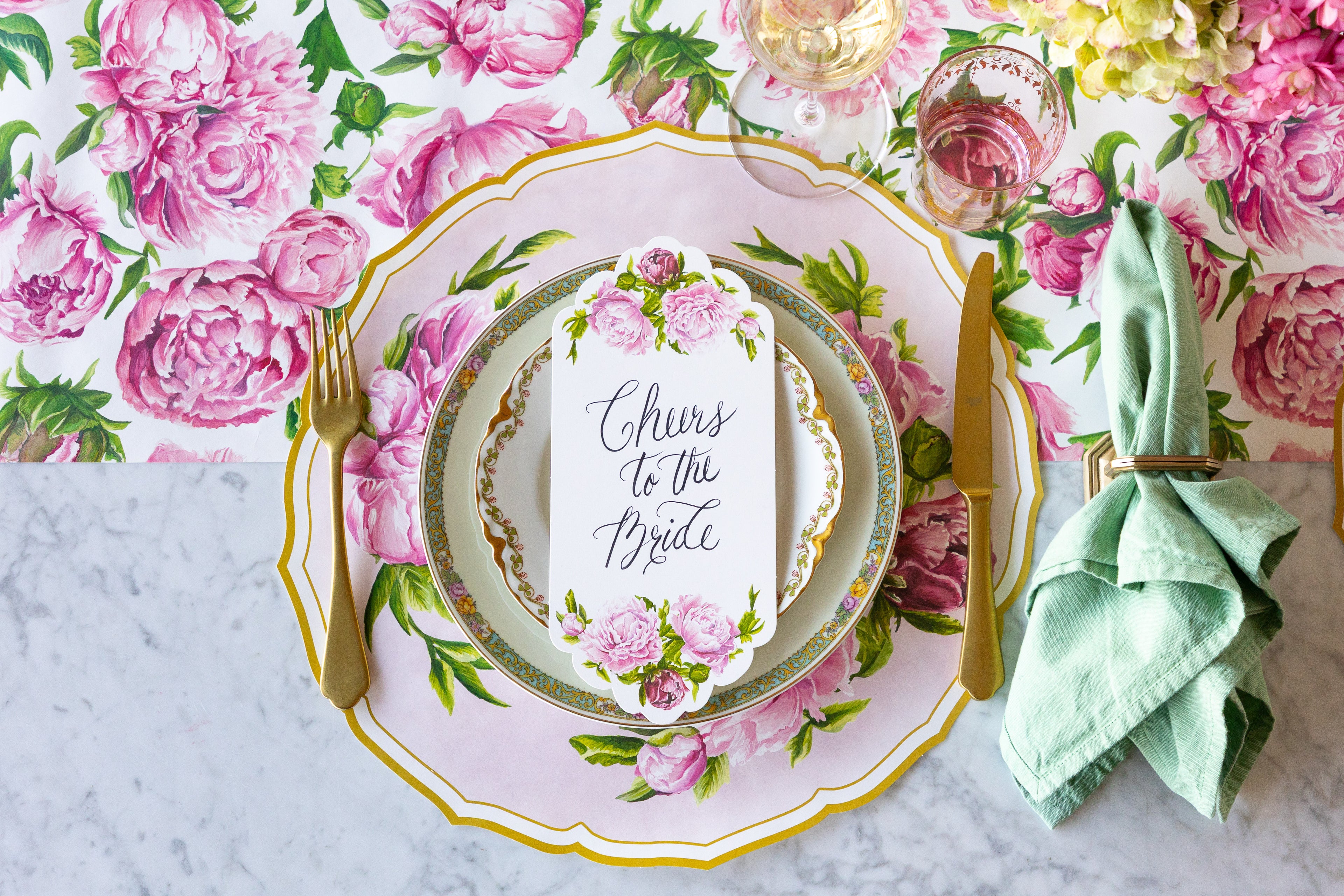 Decorative table setting with the Die-cut Peony China Placemat, floral plates, gold flatware and Peonies Table Card with "Cheers to the Bride" on the Peonies in Bloom Runner.