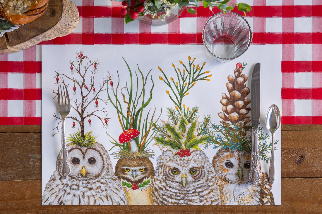 A placemat with four owls on it drawn by Vicki Sawyer.  All of the owls have festive hats on made of branches and various greenery.  All are set on a wood table with a red check table runner.
