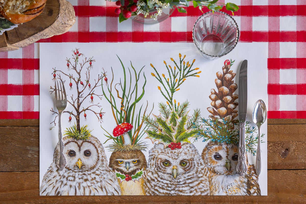 A placemat with four owls on it drawn by Vicki Sawyer.  All of the owls have festive hats on made of branches and various greenery.  All are set on a wood table with a red check table runner.