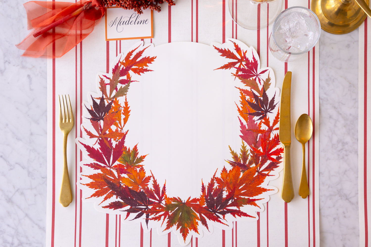 Die-cut Japanese Maple Wreath Placemat on a red striped table runner with gold cutlery and a glass. 