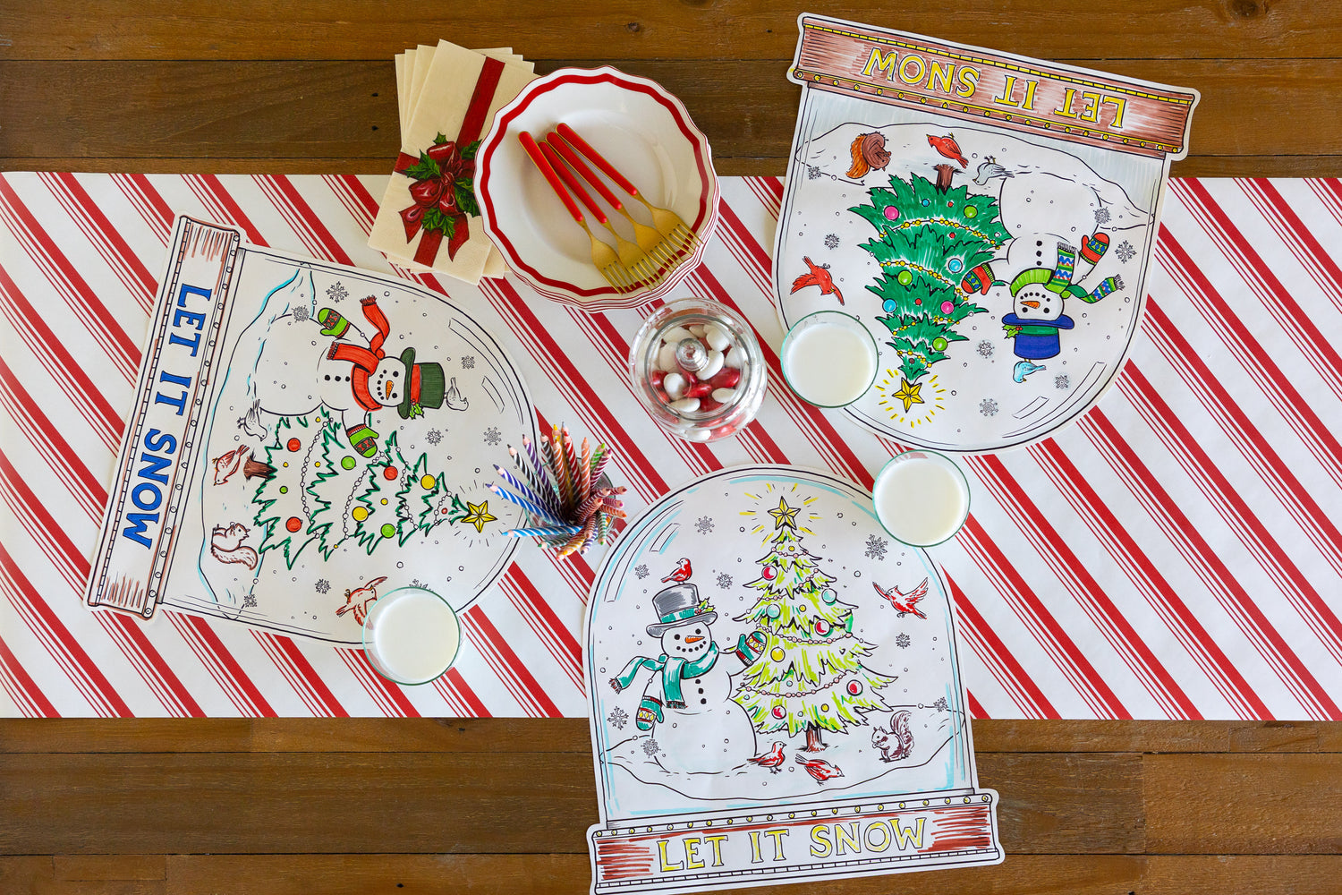Table with three partially colored in Die-cut Snow Globe Coloring Placemats on a Candy Stripe Runner with glasses of milk and candy on the table. 