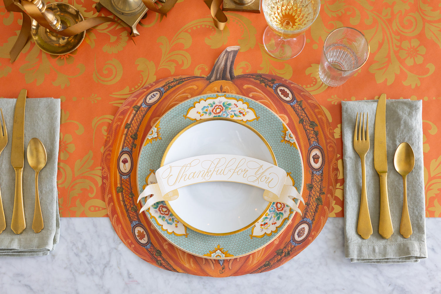 Thanksgiving table setting with pumpkin-themed placemat, gold cutlery, and decorative glasses.