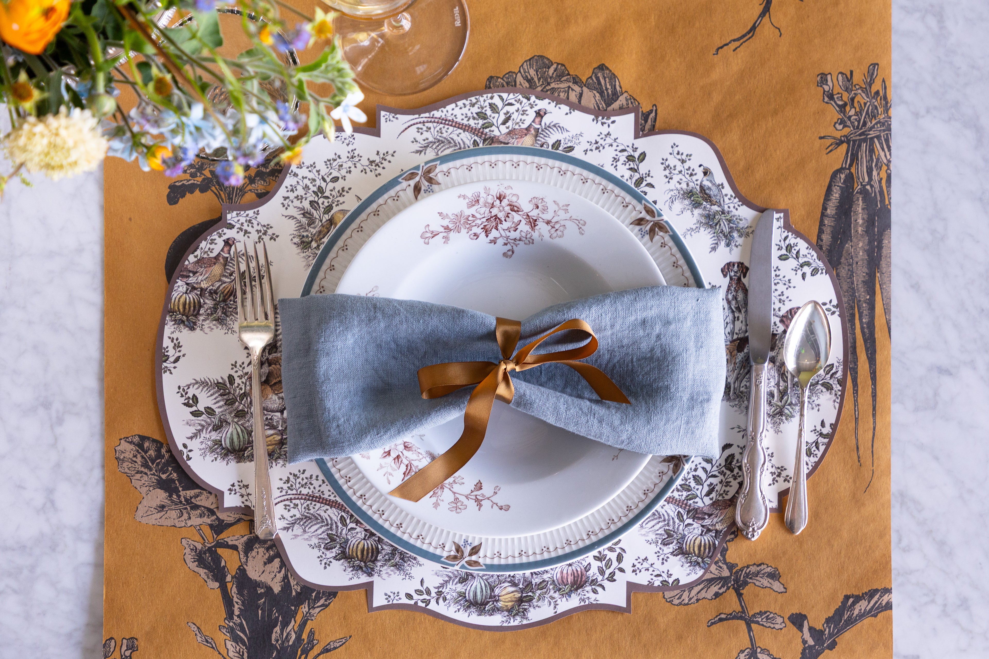 Decorative table setting with floral arrangement, plates, and cutlery on a patterned placemat.