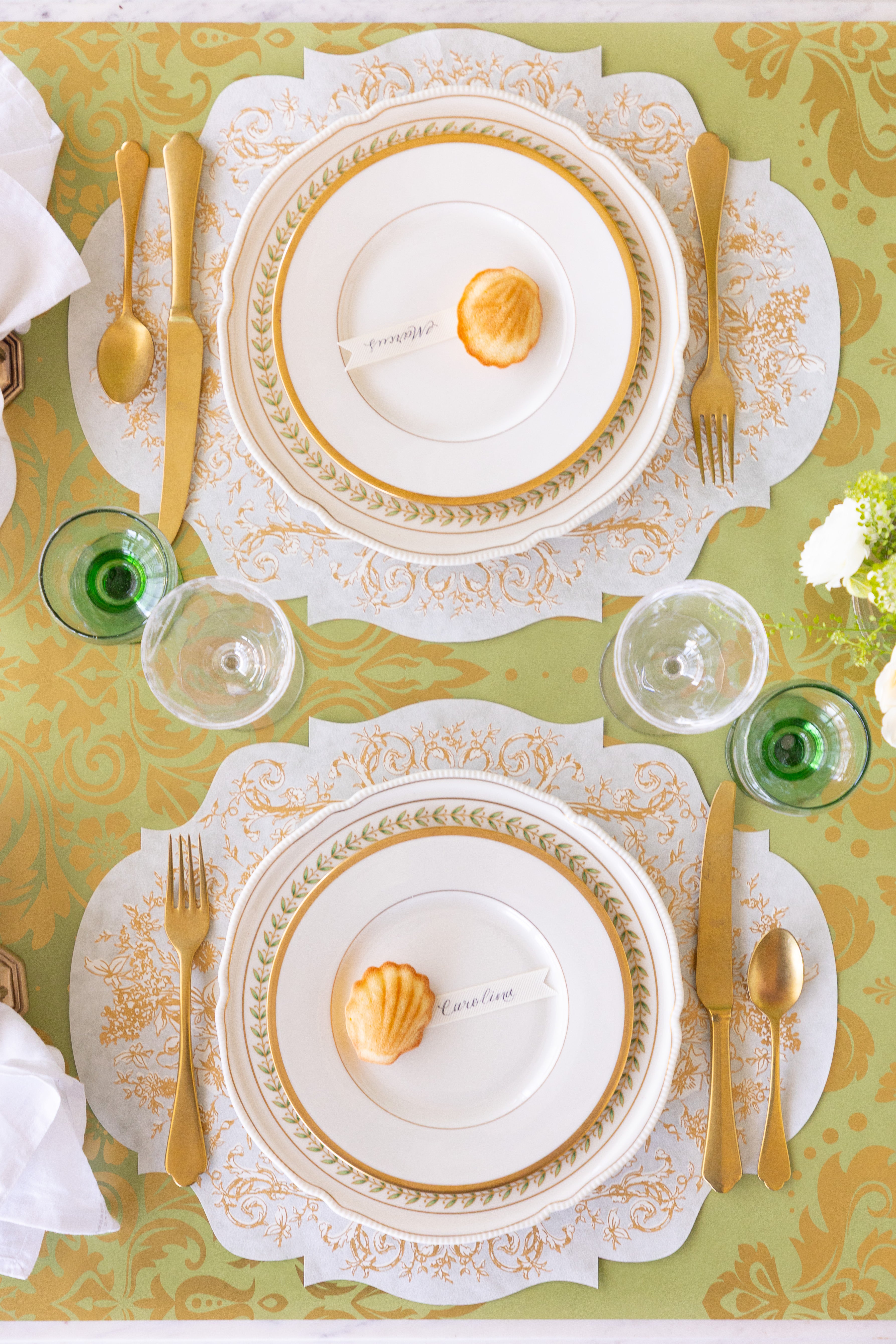 Elegant table setting with gold and white plates, cutlery, and decorative elements on a green patterned tablecloth.